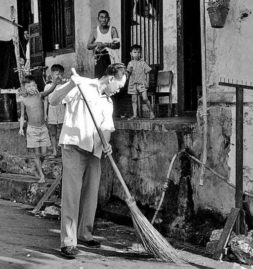 The image depicts a historical scene with a group of people outside a building. The focus is on a man sweeping the street with a large broom. The hint suggests that this man is Lee Kuan Yew. He is dressed in a light-colored shirt and pants, concentrating on the task at hand. Around him are several children and an adult watching from the steps of the building. The children are casually dressed, appearing curious or entertained by the activity. The setting appears to be an urban residential area, likely dating back to the mid-20th century, judging by the architecture and clothing styles. The image is in black and white, enhancing its historical feel. Pipes and other communal infrastructure are visible, indicating a modest neighborhood. The atmosphere is informal and communal, capturing a moment of daily life.