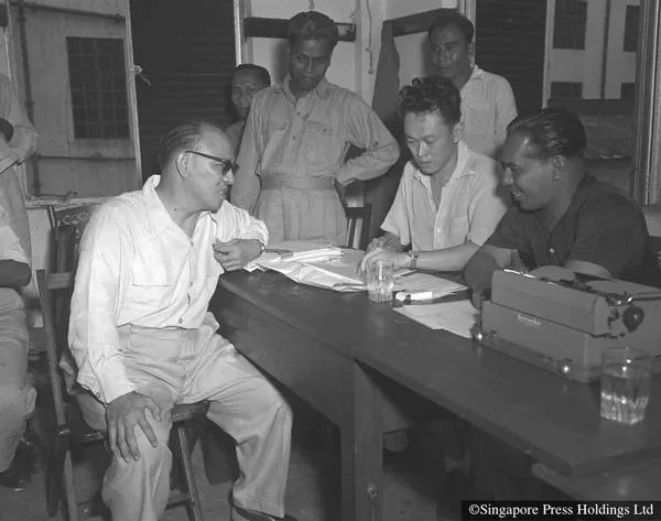 The image depicts a historical scene featuring Lee Kuan Yew, who is seated at a table engaged in discussion with several other men. The setting appears to be an office or meeting room with a simple and functional interior. Lee Kuan Yew is dressed in a light-colored shirt, intently focused on the papers in front of him. To his right, another man in a dark shirt is engaged in conversation, possibly discussing or negotiating terms. The men around the table appear involved and attentive, contributing to the sense of a serious and purposeful meeting.
In the background, other individuals stand observing or waiting for their turn to participate. The lighting is moderate, typical of indoor environments, casting soft shadows and highlighting the formal yet informal atmosphere. Notable items include a typewriter on the table, which suggests administrative work or documentation is taking place. The overall mood conveys a sense of historical importance and collaboration.
- Title: "Singapore Press Holdings Ltd"