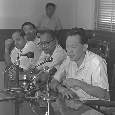 The image shows a group of four men seated at a table within a formal setting, likely a meeting or press conference. The setting is indoors with wooden paneling visible in the background and a window with blinds. The man on the far right, identified through the hint as Lee Kuan Yew, is speaking into multiple microphones placed in front of him on the table. He appears engaged and focused, suggesting he is addressing a serious topic. The other three men are seated beside him, listening attentively. They are all wearing light-colored, short-sleeved shirts, which gives a slightly casual yet professional appearance suitable for a warm climate.
The mood in the room appears serious and attentive, with each person focused on the discussion at hand. The microphones, along with the posture and expressions of the individuals, indicate that this is an important event, possibly a press conference or a political meeting. The image is in black and white, adding a historical feel to the scene, and there are no visible logos or text in the image.