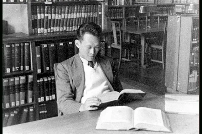 In this black-and-white photograph, Lee Kuan Yew is seated at a wooden table in a library setting, deeply engrossed in reading a book. Shelves filled with numerous books are visible in the background, contributing to the scholarly atmosphere. Lee Kuan Yew is dressed in a formal attire consisting of a suit jacket over a white sweater, projecting a sense of academic diligence. His focused posture suggests he is absorbed in his reading, embodying a moment of quiet concentration. The library appears to be well-organized, with additional seating and a lamp visible further back, indicating a space designed for study and reflection. The monochrome tone of the image adds a vintage, historical feel, highlighting a past era. The composition centers around Lee Kuan Yew, framed by the structured lines of the bookshelves, drawing the viewer’s attention to his intellectual engagement in this serene environment.