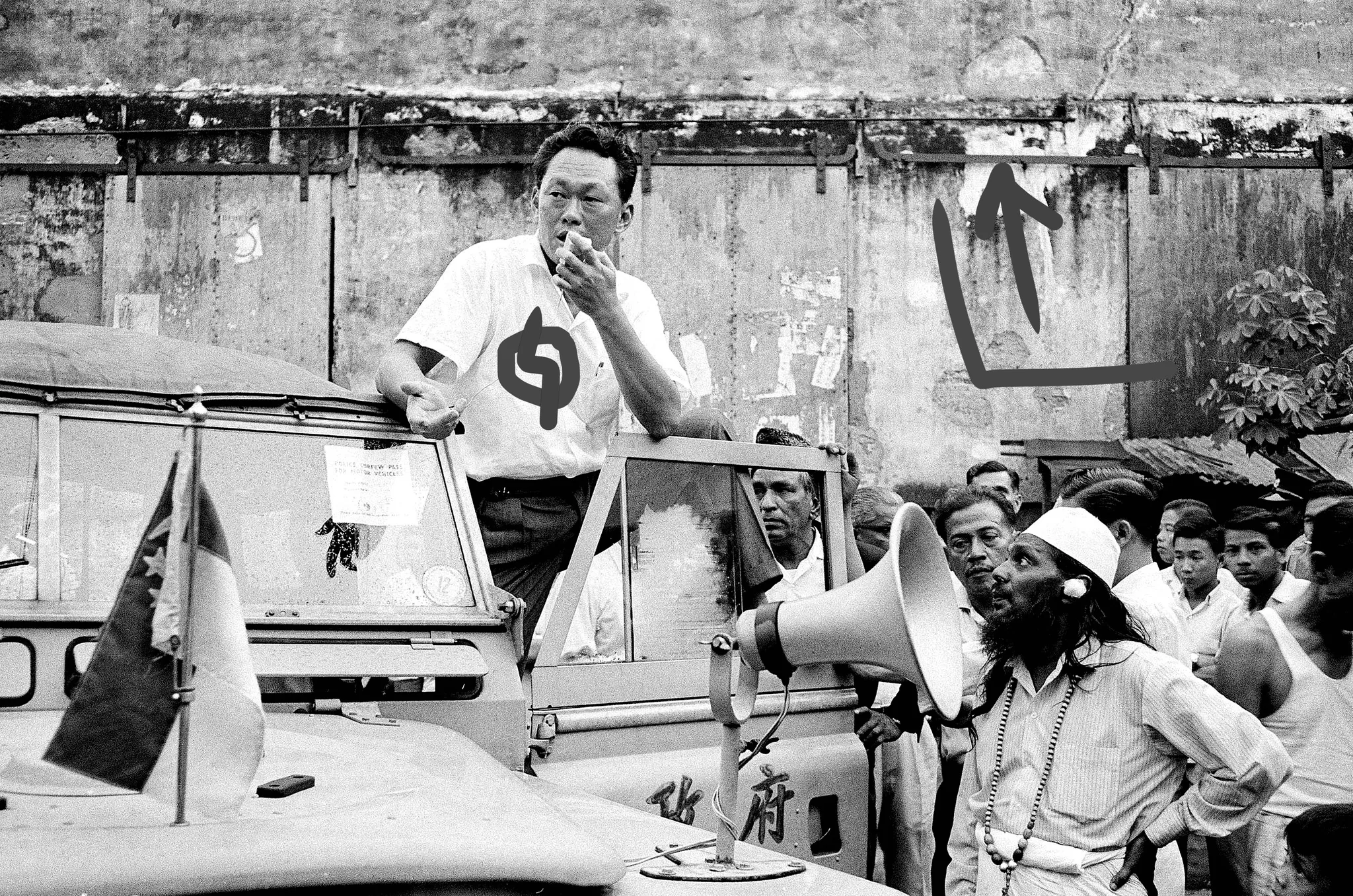 The image captures a historical scene featuring Lee Kuan Yew, as identified from the hint. It is a black-and-white photograph, likely from the mid-20th century, depicting Lee Kuan Yew standing on a vehicle, speaking to a gathered crowd. He is dressed in a light-colored short-sleeved shirt with a logo on it and appears to be addressing the public with a serious expression.
The setting is outdoors, with a backdrop of a weathered wall, suggesting an urban environment. A group of people surrounds him, including a man with a beard and a cap, who is holding a loudspeaker. The vehicle has several items on it, including flags and a sign that reads "POLICE ENTRY PASS TO THE VEHICLE." The crowd consists of men in various attire, some appearing intent on listening while others seem more relaxed. The overall mood of the photograph is tense and focused, capturing a moment of political significance. The black-and-white style adds a timeless and historical quality to the scene.