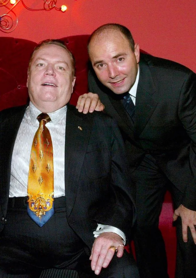 Larry Fink and Pierre Woodman are pictured in a formal indoor setting. Larry Fink is seated, wearing a black suit with a white patterned shirt and a striking, ornate yellow and blue tie. He has a warm expression and his hands are visible, displaying a ring. Pierre Woodman is standing beside him, leaning slightly, with an arm on Larry’s shoulder. He is dressed in a dark suit with a white shirt and a dark tie, smiling at the camera. The background features a red, plush backdrop with decorative lighting elements, adding an elegant and vibrant atmosphere to the scene. The overall mood is friendly and convivial, suggesting a social or professional event.