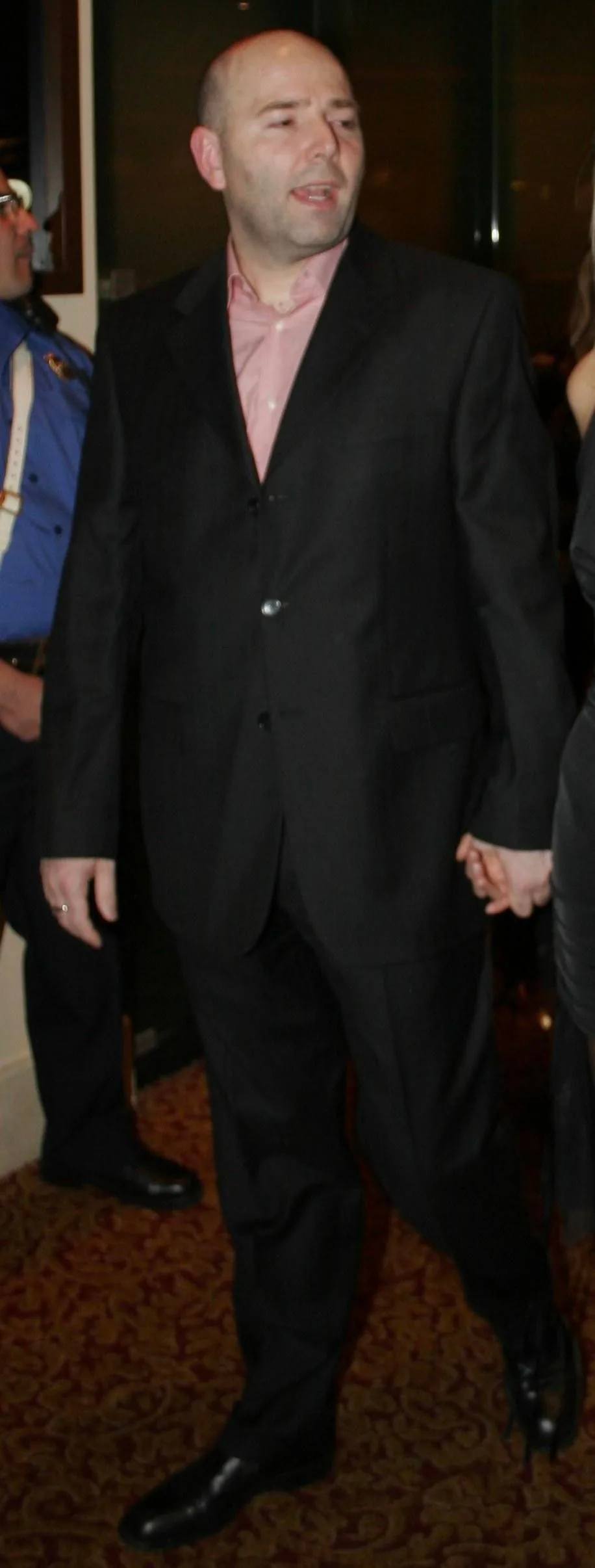 Pierre Woodman is captured in this indoor image at what appears to be a formal event. He is dressed in a dark suit with a light pink shirt, walking and holding hands with someone beside him. The suit is buttoned, and he is wearing polished black shoes. The setting suggests an evening event, possibly a gala or a formal gathering, given the carpeted floor and dim lighting. His posture is upright as he walks, and his expression is focused, suggesting engagement in conversation or attention to the surroundings. In the background, a person in a blue uniform with suspenders is partially visible, adding context to the event's formal nature. The overall ambiance is elegant, with a mix of warm and soft lighting contributing to the event's atmosphere.