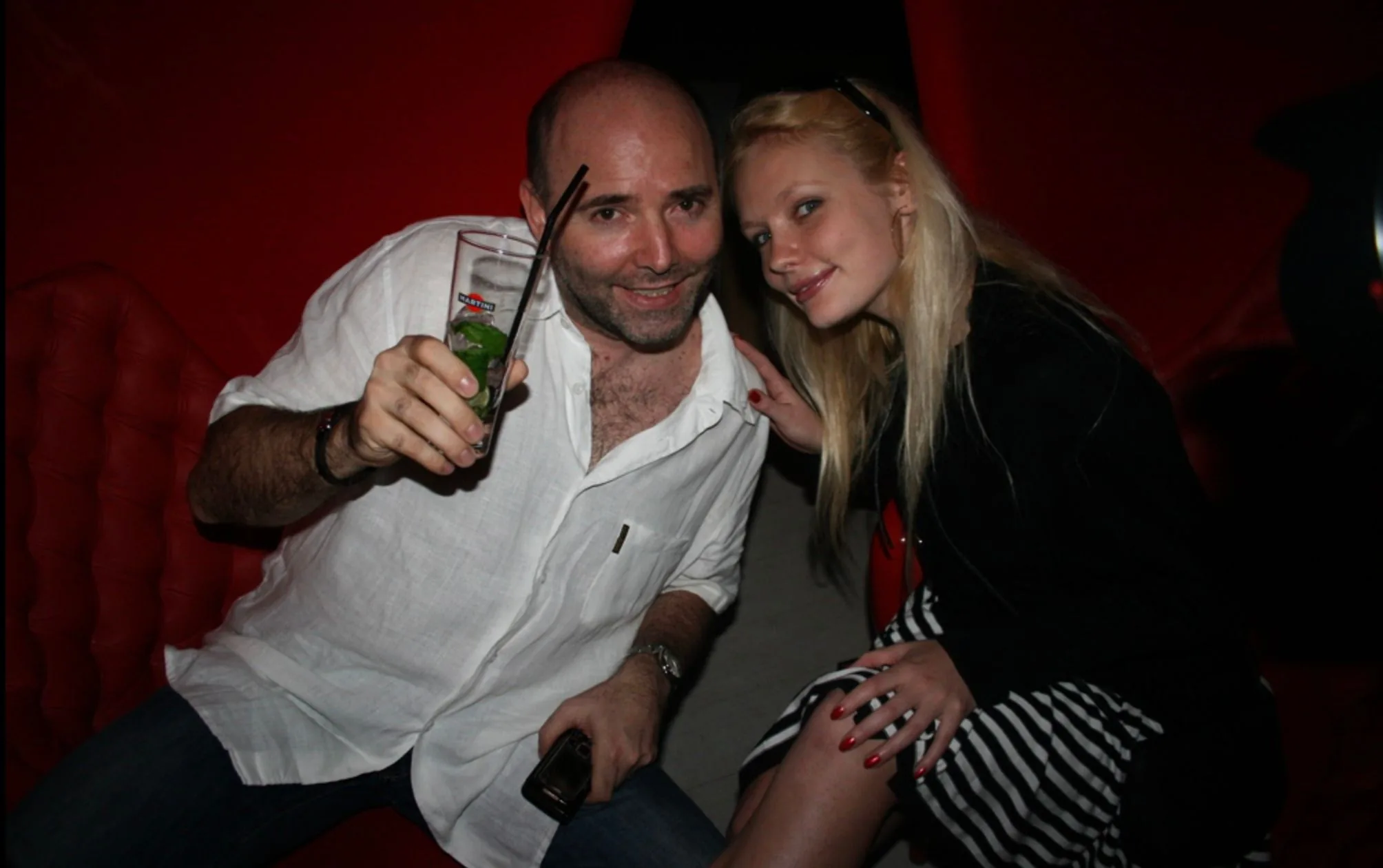 The image captures a lively social scene featuring two individuals seated in a dimly lit, indoor setting. The person on the left, identified as Pierre Woodman, is dressed in a casual white shirt and dark jeans. He holds a tall glass with the word "MARTINI" visible on it, containing a cocktail garnished with mint leaves and a black straw. His demeanor is cheerful, and he appears to be engaging with the camera directly, exuding a relaxed and jovial vibe.
Beside him, a woman with long blonde hair and a friendly smile leans slightly towards him. She is wearing a black top paired with a striped skirt, and her red nails add a pop of color to the scene. Both individuals seem to be enjoying the moment, suggesting a convivial atmosphere typical of a party or social gathering.
The background features red upholstered seating, which complements the overall warm and cozy ambiance of the setting. The lighting is soft, emphasizing the intimate and friendly nature of the encounter. The focus is primarily on the two individuals, with the rest of the setting mostly blurred out.