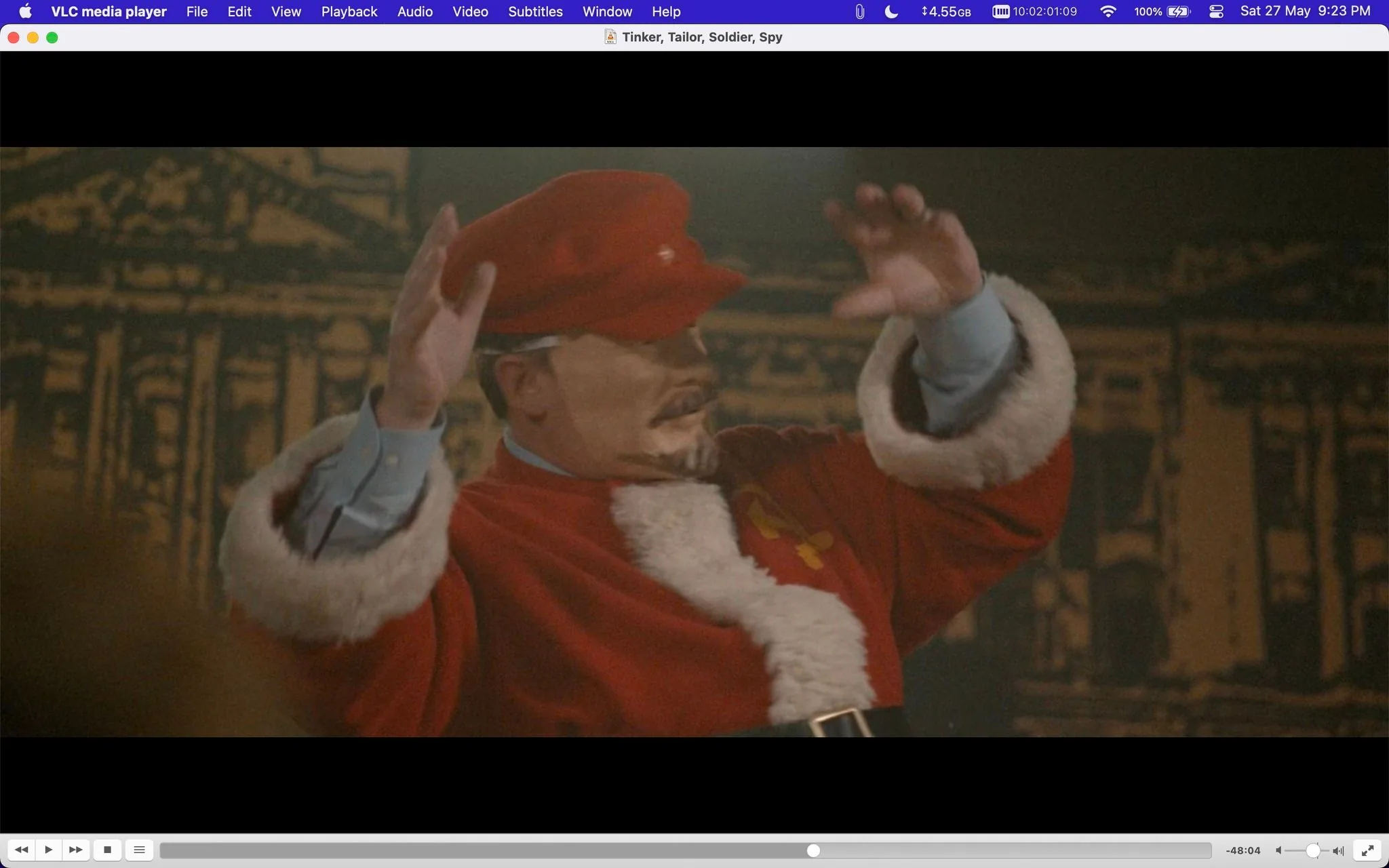 The image is a screenshot from a scene in the film "Tinker, Tailor, Soldier, Spy," displayed on VLC media player. It shows a person dressed in a red and white costume reminiscent of a mix between Santa Claus and a Soviet-style uniform, including a red hat and a coat with fur trim. The figure appears to be performing, with their hands raised, suggesting they are in the midst of an expressive action. The background features a dark, possibly theatrical backdrop with architectural motifs, adding to the dramatic setting. The lighting is somewhat subdued, evoking a stage or dimly lit environment. The scene conveys a mix of theatricality and intrigue, fitting the film’s espionage theme. There is no visible text or subtitles in this frame.