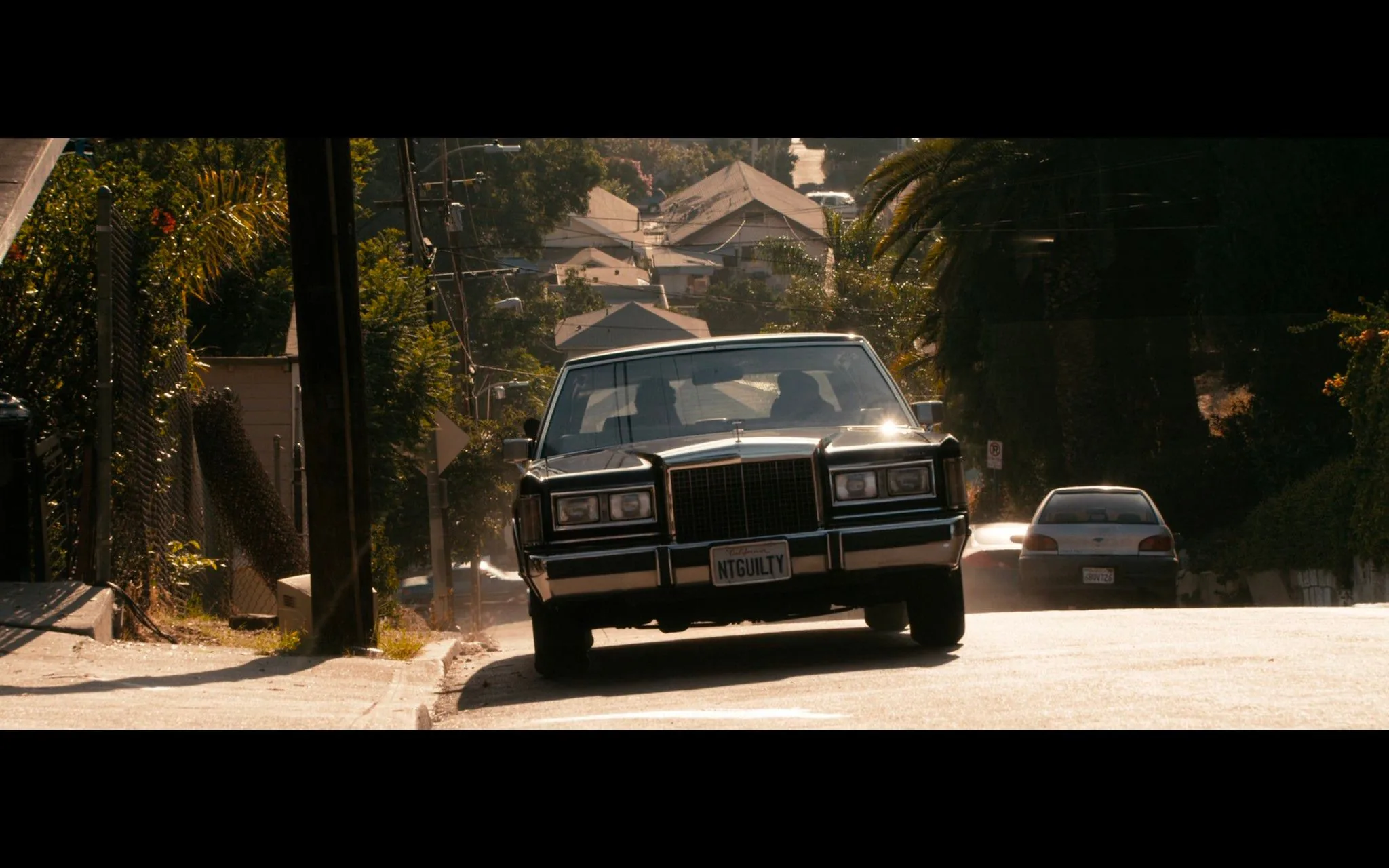 ```
In this scene from "Lincoln Lawyer," a classic black Lincoln Town Car is prominently featured, driving up a residential street during the day. The vehicle's distinctive vanity license plate reads "NTGUILTY," adding a layer of intrigue and character relevance. The car occupies the center of the frame, with its headlights and chrome detailing catching the sunlight, creating a striking visual against the urban backdrop. The background is composed of suburban houses and lush greenery, providing a contrast to the sleek, polished appearance of the car. The lighting is warm and natural, suggesting a sunny day that enhances the vividness of the scene. The image captures a moment of movement and purpose, likely reflecting the film's themes of justice and legal maneuvering. The setting is unmistakably urban, with power lines and palm trees lining the street, adding to the Hollywood ambiance.