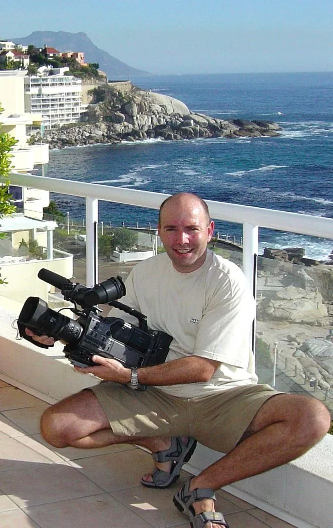 The image features Pierre Woodman, who is crouching on a balcony with a scenic coastal view in the background. He is holding a professional video camera, suggesting his involvement in videography or filmmaking. The background showcases a picturesque ocean scene with rocky cliffs and buildings along the coastline, likely a beachside location. The sky is clear, indicating a bright, sunny day. Pierre is casually dressed in a light-colored t-shirt, shorts, and sandals, appearing relaxed and comfortable. His expression is cheerful, suggesting he is enjoying the moment or the work he is engaged in. The composition captures both the technical aspect of filmmaking and the serene beauty of the coastal environment, presenting a harmonious blend of professional and leisurely elements. The lighting highlights the vibrancy of the setting, enhancing the visual appeal of both Pierre and the surrounding landscape.