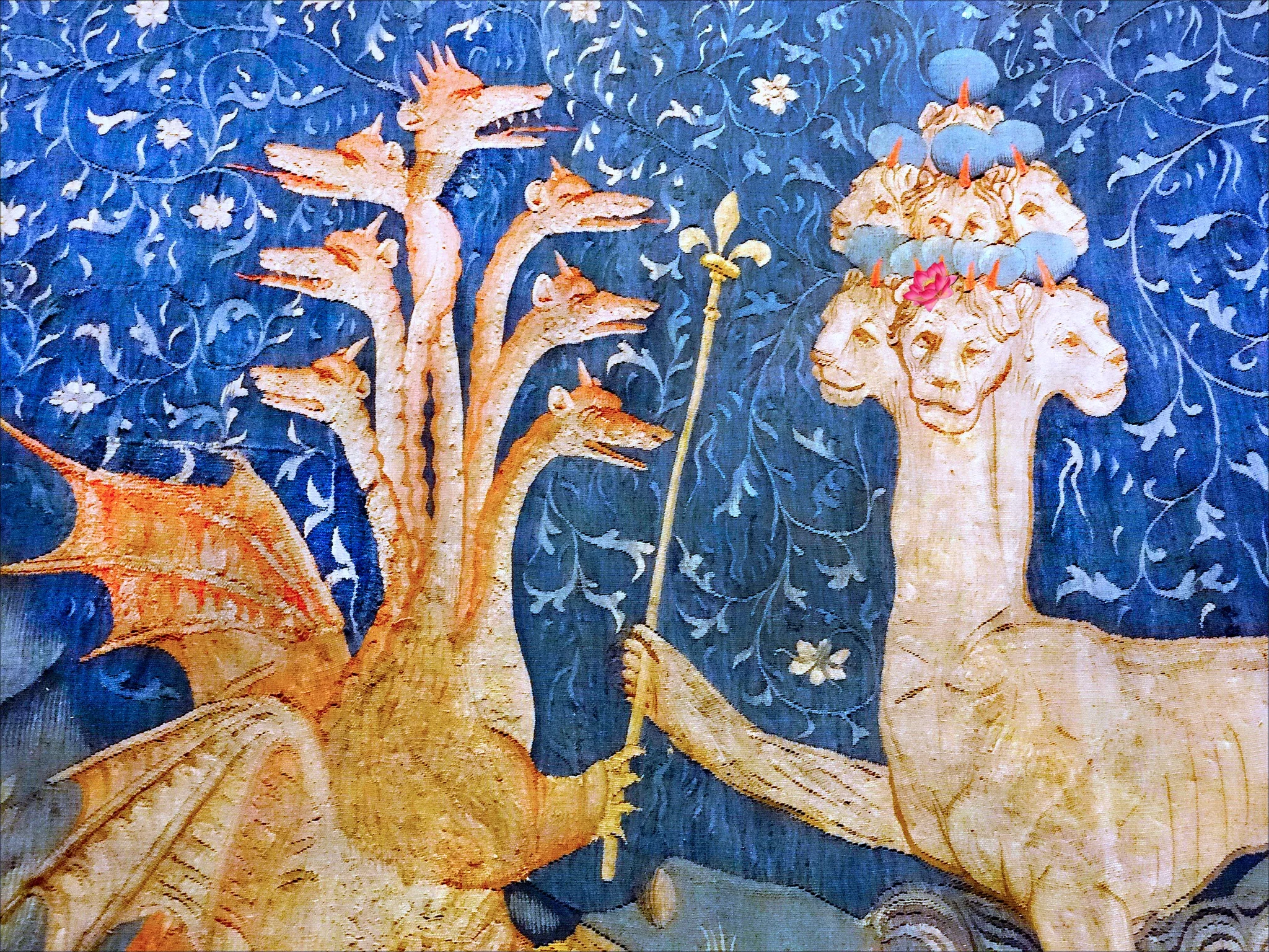 This image is an illustration from "The Apocalypse Tapestry," a famous medieval tapestry depicting scenes from the Book of Revelation. In the foreground, there is a dramatic confrontation between two mythological creatures. On the left, a multi-headed dragon, each head with an open mouth, showcases its impressive wings, colored in shades of orange and gold. The dragon appears lively and dynamic, suggesting movement and fierceness. On the right, another fantastical beast with multiple crowned heads is depicted. The heads are crowned with blue and adorned with a single pink flower atop. This creature holds a scepter, adding a regal and authoritative aura.
The background is a rich blue, filled with intricate vine and floral patterns, adding depth and elegance to the scene. The tapestry style is characteristic of medieval art, using bold colors and detailed embroidery to convey a narrative. The overall mood is one of conflict and grandeur, characteristic of the apocalyptic themes in the tapestry. The craftsmanship highlights the skill and artistry of medieval weavers, presenting a vivid interpretation of a biblical story.