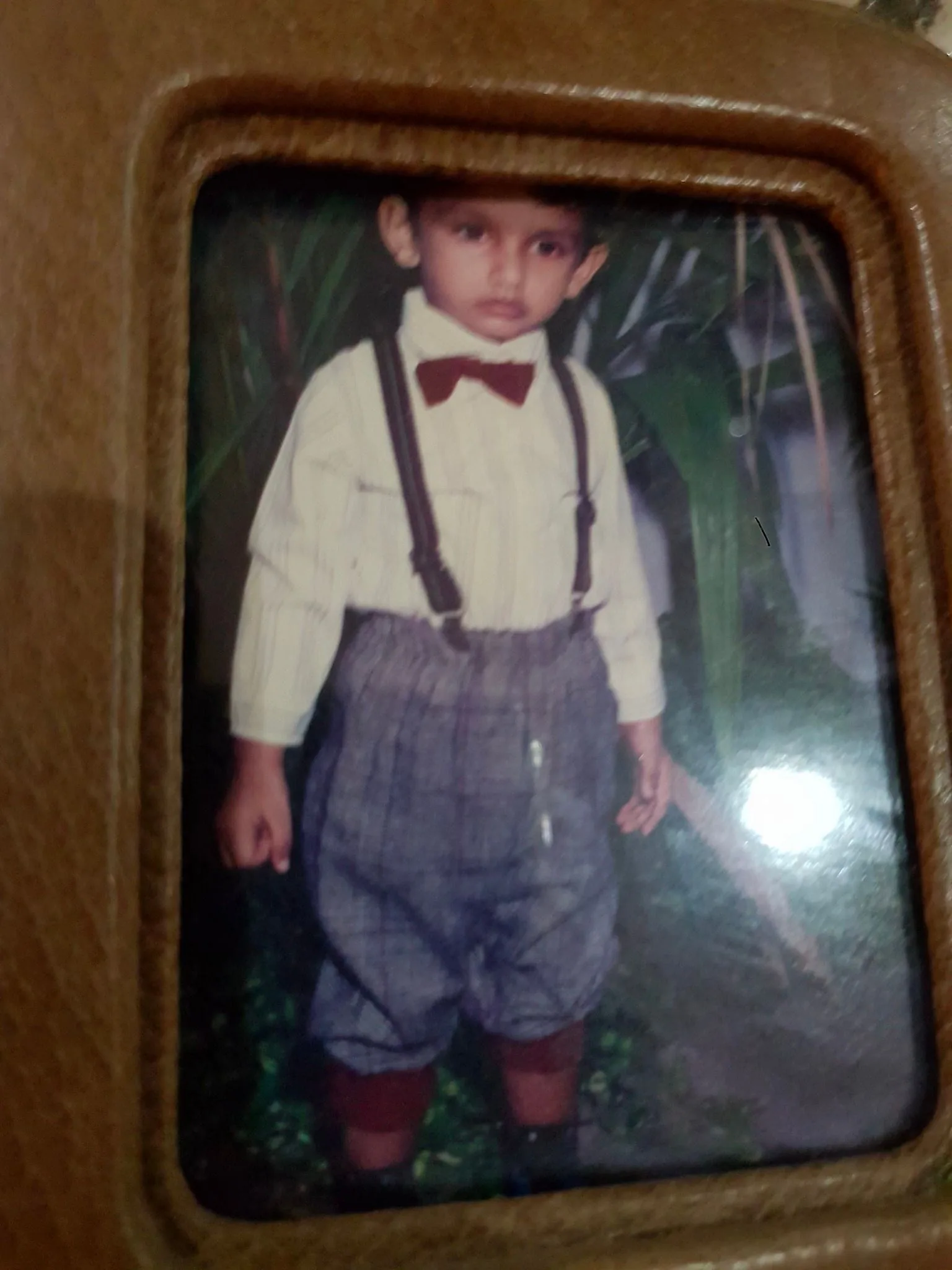Lehan Edirisinghe is depicted as a young child in this charming vintage photograph. He is dressed in a formal outfit that includes a striped shirt with a dark red bow tie, giving him a classic and sophisticated look. His suspenders are neatly fastened, and he wears high-waisted plaid shorts, adding a touch of old-fashioned elegance. The shorts are complemented by knee-high socks, which are slightly visible above his shoes, completing the formal attire. Lehan stands in a garden-like setting, with greenery subtly visible in the background, enhancing the natural and serene atmosphere of the photo. His expression is calm and composed, adding to the overall charm of the image. The photograph is encased in a wooden frame, which suggests it might be a treasured family keepsake. The lighting is soft, highlighting the nostalgic feel of the photograph, capturing a moment of childhood innocence and style.