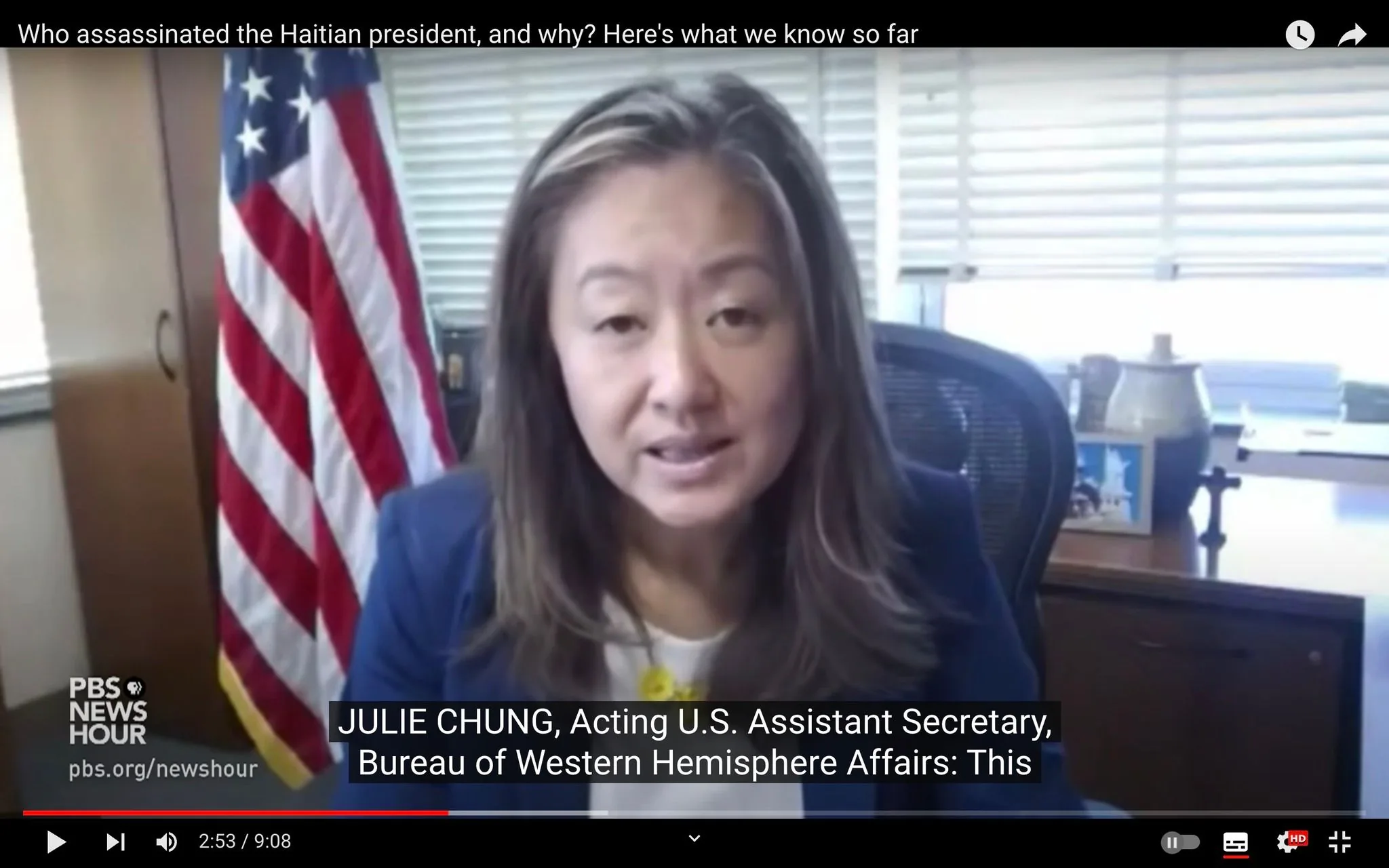 The image is a video frame from a YouTube video titled "Who assassinated the Haitian president, and why? Here's what we know so far" on the PBS NewsHour channel. In the frame, Julie Chung, identified as the Acting U.S. Assistant Secretary for the Bureau of Western Hemisphere Affairs, is speaking during an interview. She is seated in an office setting with an American flag prominently displayed behind her, suggesting a formal government environment.
Julie Chung is wearing a professional outfit, with a navy blazer over a white top. Her expression is earnest, reflecting the serious nature of the subject matter. There is a wooden desk in the foreground with various items, including a framed photograph and a decorative vase. The lighting is neutral, giving the scene a clear and professional appearance.
The on-screen text includes the PBS NewsHour logo and website, along with subtitles that capture Julie Chung's role and her speech. The video discusses the assassination of the Haitian president, a topic of significant geopolitical interest.
