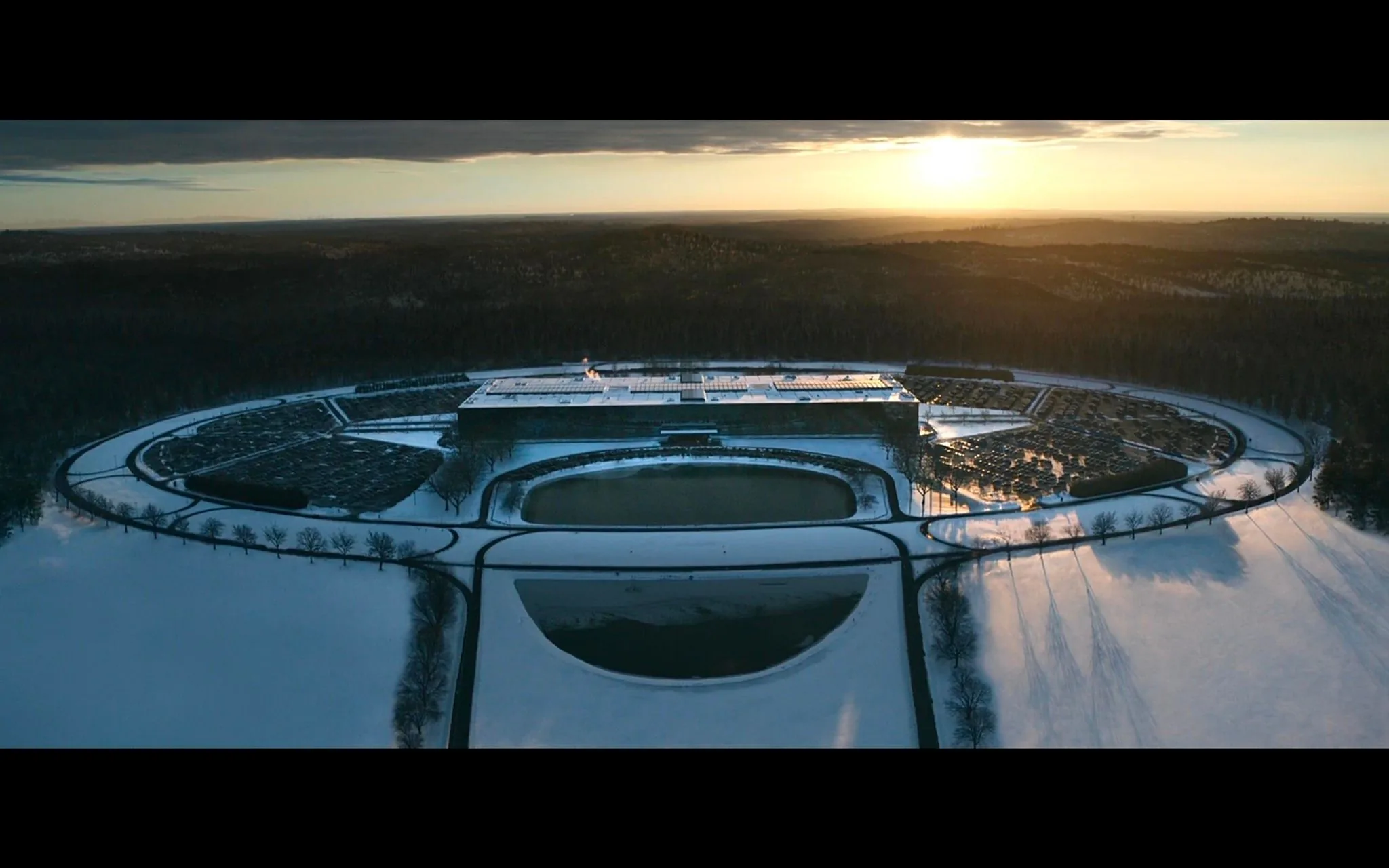This image is an aerial view of a large, modern architectural structure surrounded by snowy fields. The building is centrally positioned within a circular arrangement of roads and pathways, giving it a futuristic appearance. The photograph is taken at sunset, casting long shadows across the snow-covered landscape and adding a warm glow to the scene. The structure seems to include large parking areas filled with cars, suggesting it's a facility or corporate complex. The presence of a body of water in front of the building adds to the symmetry and visual appeal. The landscape in the background is a vast expanse of forested area, indicating a remote location. The overall mood is serene and quiet, with the winter setting enhancing the stillness. There is no visible text or specific identifying features in the image that suggest a particular site or app.