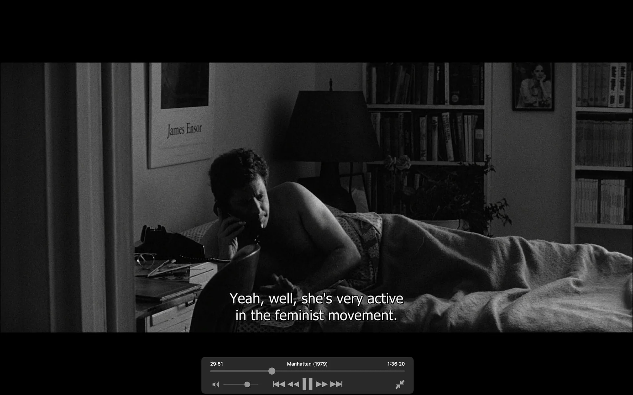 The image is a black-and-white video frame from the film "Manhattan" (1979). It depicts a man lying in bed, partially covered by a blanket, while talking on a corded phone. The lighting is dim, suggesting a nighttime setting, with the lamp on the bedside table turned off. The room has a cozy, lived-in feel, with a bookshelf filled with books visible in the background. There's a framed poster on the wall with the name "James Ensor" visible, adding an artistic touch to the room. The man appears to be engaged in a late-night conversation, as indicated by the caption, "Yeah, well, she's very active in the feminist movement." The scene captures a moment of introspection and communication, typical of the film's thematic exploration of personal relationships.