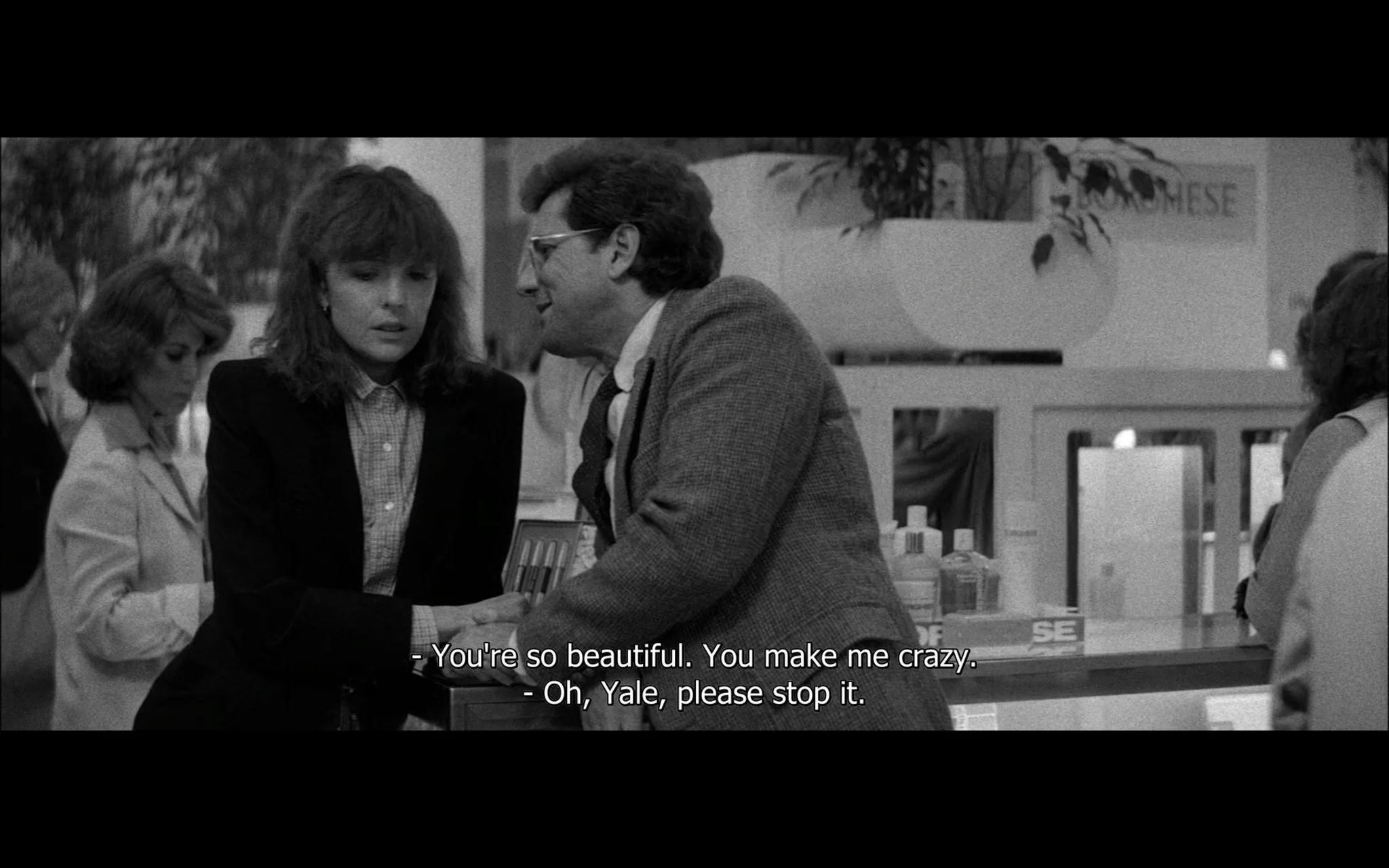 This black-and-white video frame is from the film "Manhattan." The scene takes place in a department store, characterized by its indoor setting with cosmetic products and displays visible in the background. The image features two main characters engaged in a conversation. The man, referred to as Yale, is leaning towards the woman in a gesture that suggests intimacy or persuasion. His attire consists of a suit jacket and glasses, adding to his scholarly appearance. The woman, appearing somewhat uncomfortable, looks away from Yale. She is dressed in a formal, buttoned-up outfit, and her expression suggests reluctance or unease, in line with her dialogue telling Yale to stop.
In the background, other shoppers can be seen, contributing to the bustling atmosphere typical of a department store. The lighting is even, highlighting the emotional tension between the characters. The dialogue in the subtitles reveals a personal and possibly romantic dynamic, with Yale expressing admiration and the woman resisting his advances. This scene captures a moment of complex interpersonal interaction within the sophisticated, urban backdrop of the film.