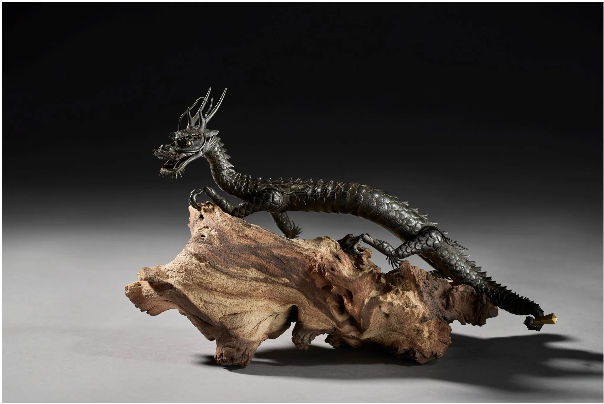 The image features a detailed sculpture of a dragon, artfully crafted to stand on a piece of driftwood or similarly textured wood. The dragon is intricately designed with a long, sinuous body covered in scales, which gleam under the lighting, highlighting the artistry of the piece. Its head is fierce, with sharp horns and a snarling expression, capturing a sense of movement and life as it appears to crawl over the wood beneath it. The dragon's claws are extended, gripping the wood as if in motion. The sculpture is set against a neutral, dark background, which contrasts with the lighter hues of the wood and the metallic sheen of the dragon, emphasizing the creature's form and details. The lighting is subtle yet effective, casting shadows that add depth and drama to the scene, enhancing the visual impact of the dragon's dynamic pose. The overall composition suggests a theme of mythical or fantastical art, focusing on the elegance and power of the dragon motif.
