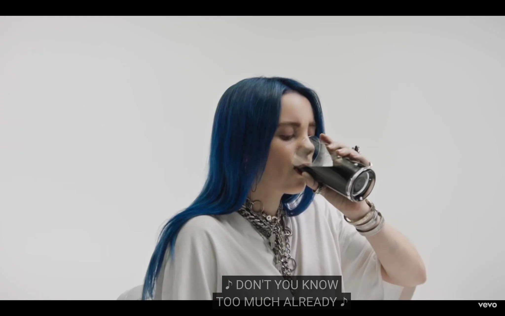 The image is a frame from a music video featuring Billie Eilish. She is seated indoors, drinking from a glass filled with a dark liquid, possibly soda or coffee. Billie has long, blue hair and is wearing a casual white outfit accented with a chunky silver chain necklace and multiple bracelets. Her eyes are closed as she takes a sip, suggesting calmness or introspection. The background is simple and white, focusing attention on her. Captions at the bottom of the frame read, "DON'T YOU KNOW TOO MUCH ALREADY," adding a lyrical element from the video. The lighting is clear and bright, enhancing the clean and minimalistic aesthetic typical of Billie Eilish's music videos.