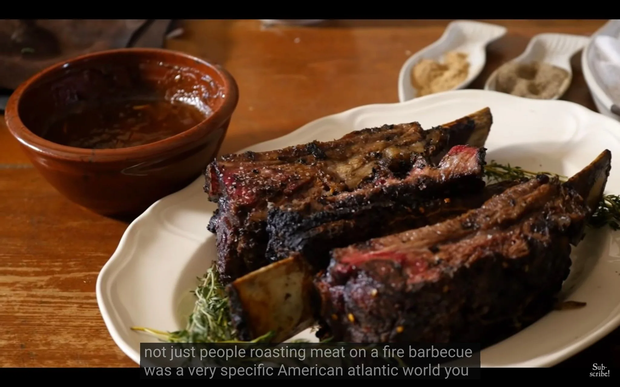 The image shows a close-up of a plate with barbecue ribs. The ribs are charred and seasoned, placed on a white dish with sprigs of herbs, likely rosemary. Next to the plate is a small brown bowl containing a sauce, possibly barbecue sauce, suggesting a setting of a meal or food preparation. In the background, there are small dishes containing various seasonings or rubs, enhancing the culinary theme. The lighting is warm, highlighting the texture and color of the meat. The table appears wooden, adding to a rustic or homely atmosphere. Captions at the bottom mention the idea of barbecue in an American Atlantic context, hinting at a discussion of cultural aspects of barbecue. There are no visible titles or channel names, and the image focuses on the food presentation.