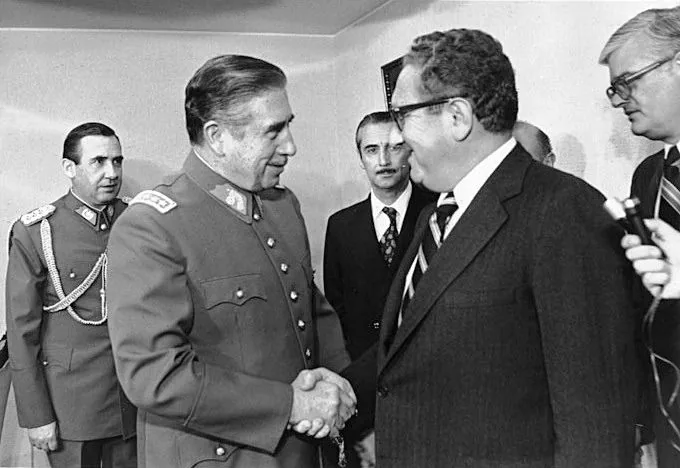In this black-and-white photograph, Henry Kissinger is prominently featured shaking hands with a man in military uniform. The setting appears to be an indoor meeting or diplomatic engagement. Henry Kissinger, wearing a suit and tie, smiles as he engages with the man in uniform, suggesting a formal yet cordial atmosphere. The man in uniform, likely a high-ranking military official given his attire, is also smiling, indicating a respectful interaction.
Several other individuals are present in the background, dressed in suits and military attire, observing the exchange. The setting is likely an office or conference room, characterized by plain walls and formal attire, which underscores the official nature of the meeting. The composition and posture of the individuals suggest a moment of politeness and diplomacy, typical of political meetings. The lighting is even, typical of indoor environments, ensuring clear visibility of the subjects.