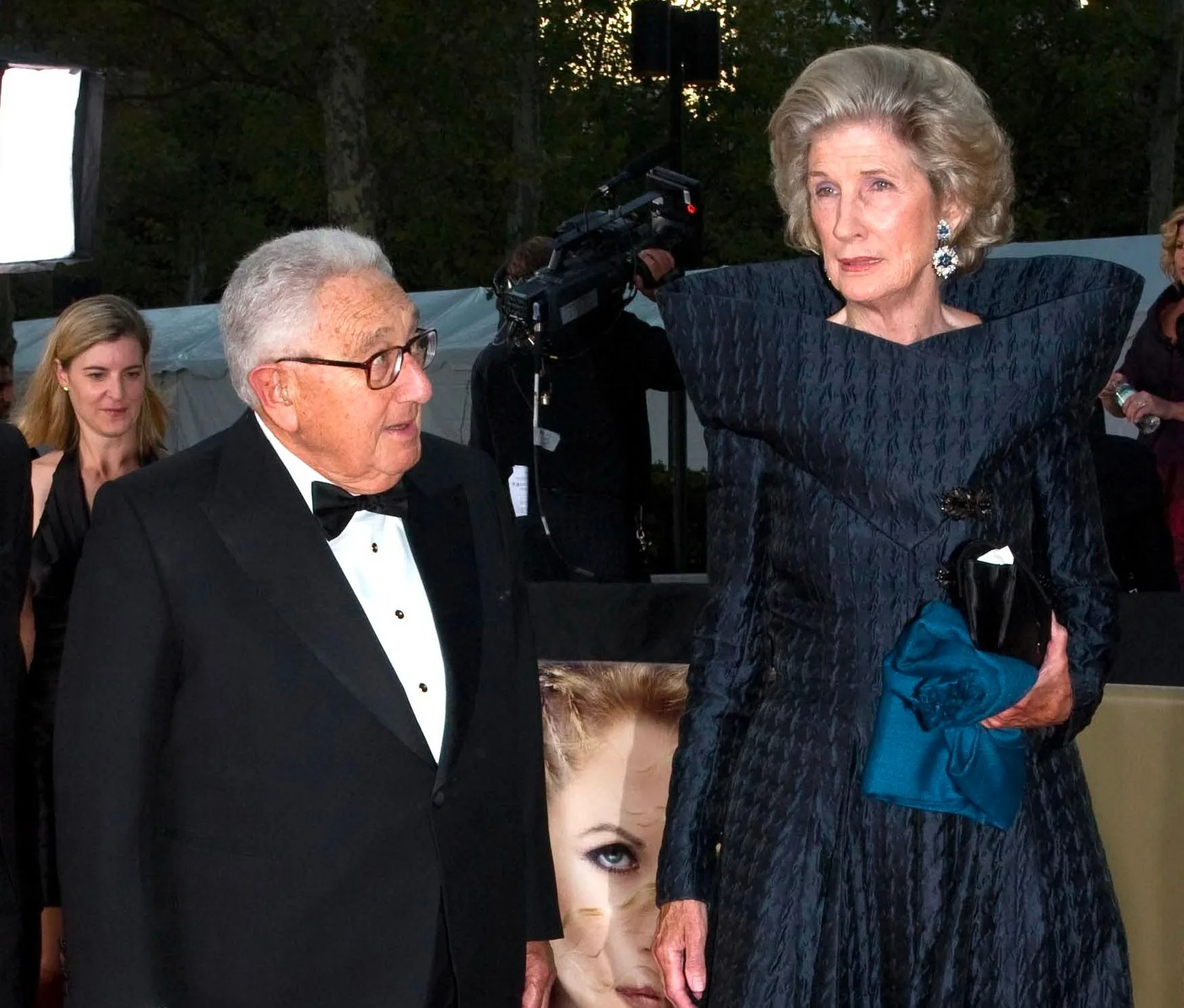 The image captures a formal event taking place outdoors, possibly in the evening due to the lighting. Henry Kissinger, identified by the user hint, is present on the left side of the image, dressed in a classic black tuxedo with a bow tie and glasses. His expression appears engaged as he looks towards the woman beside him.
The woman next to him is wearing an elegant, dark-colored gown with a distinctive high collar and textured fabric. She holds a black clutch and a blue accessory, adding a touch of color to her attire. Her hair is styled in an elegant updo, and she wears large, sparkling earrings.
In the background, there are other attendees visible, some dressed in formal attire, and a large camera setup suggests media coverage. The area is surrounded by trees and a white tent, hinting at a high-profile event. The overall mood is sophisticated and celebratory, with warm lighting enhancing the ambiance.