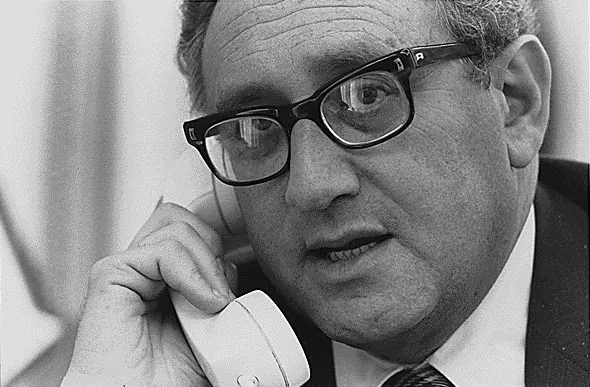 ```
Henry Kissinger is depicted in a black-and-white photograph, engaged in a phone conversation. He is holding a traditional corded telephone to his ear, suggesting a scene from a past era when such phones were commonplace. Kissinger's expression appears focused and serious, indicative of an important discussion. He wears dark-rimmed glasses, a characteristic feature, and a suit, which reflects a formal setting, likely related to his diplomatic or political role. The background is blurred, keeping the emphasis on his face and the phone, which underscores the mode of communication. The lighting is even, highlighting his facial features clearly, and the overall mood suggests a professional yet intense moment captured in time.