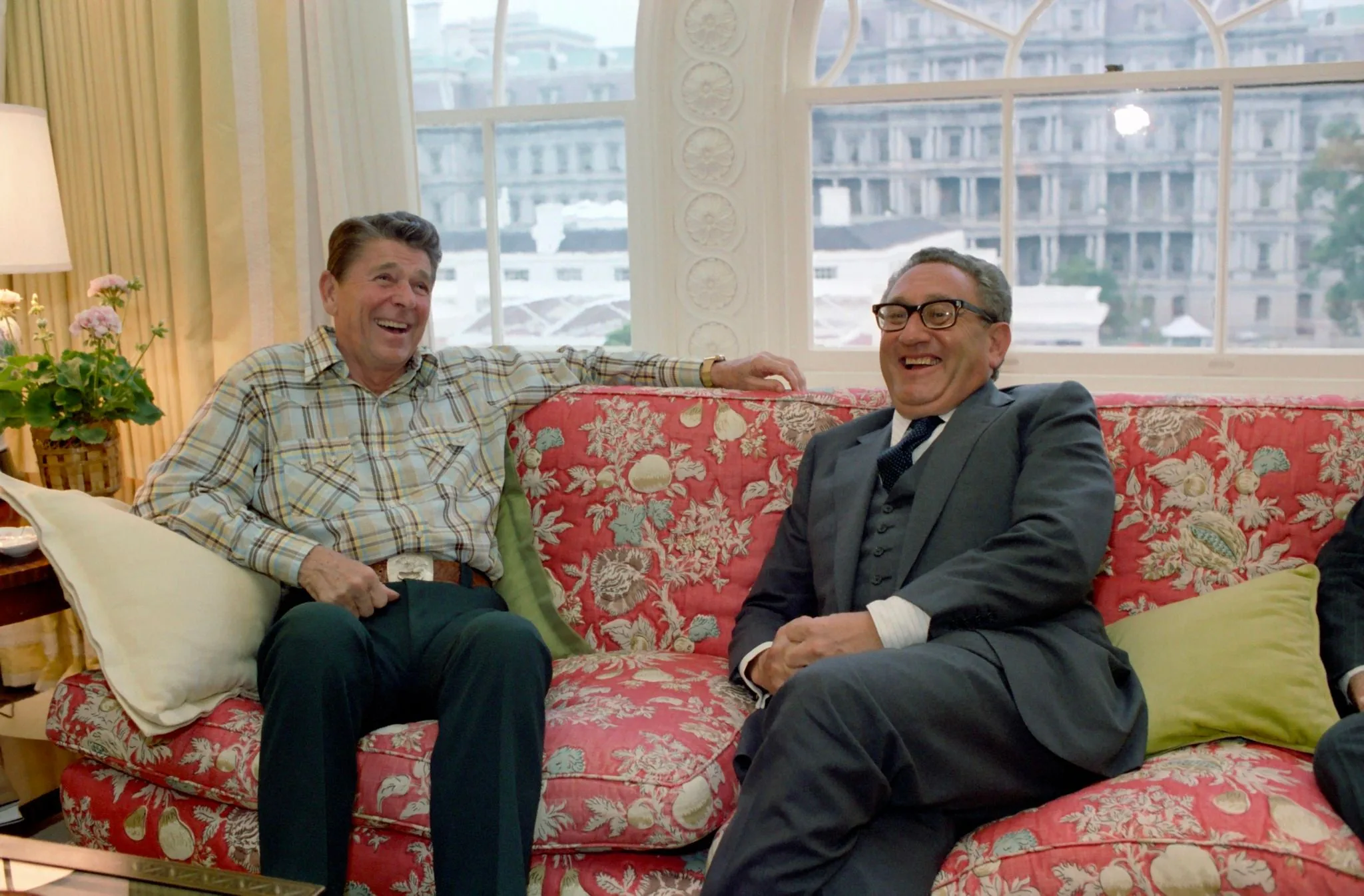 Henry Kissinger and Ronald Reagan are seated together on a floral-patterned sofa in a warmly lit room. Ronald Reagan, wearing a casual plaid shirt and dark pants, appears relaxed and jovial, leaning back comfortably with a broad smile. His demeanor is informal, indicated by his choice of attire and laid-back posture. Henry Kissinger, in contrast, is dressed in a more formal suit, complete with a tie and glasses. He is also smiling, contributing to the friendly atmosphere of the scene.
The room is tastefully decorated, with cream-colored curtains and a lamp with a soft glow on a side table. A potted plant adds a touch of greenery to the setting. Behind them, a large window offers a view of an ornate building outside, enhancing the elegance of the location. The overall atmosphere is one of camaraderie and ease, suggesting a moment of shared humor or pleasant conversation between the two figures. The lighting is bright and natural, emphasizing the cheerful mood of the meeting.