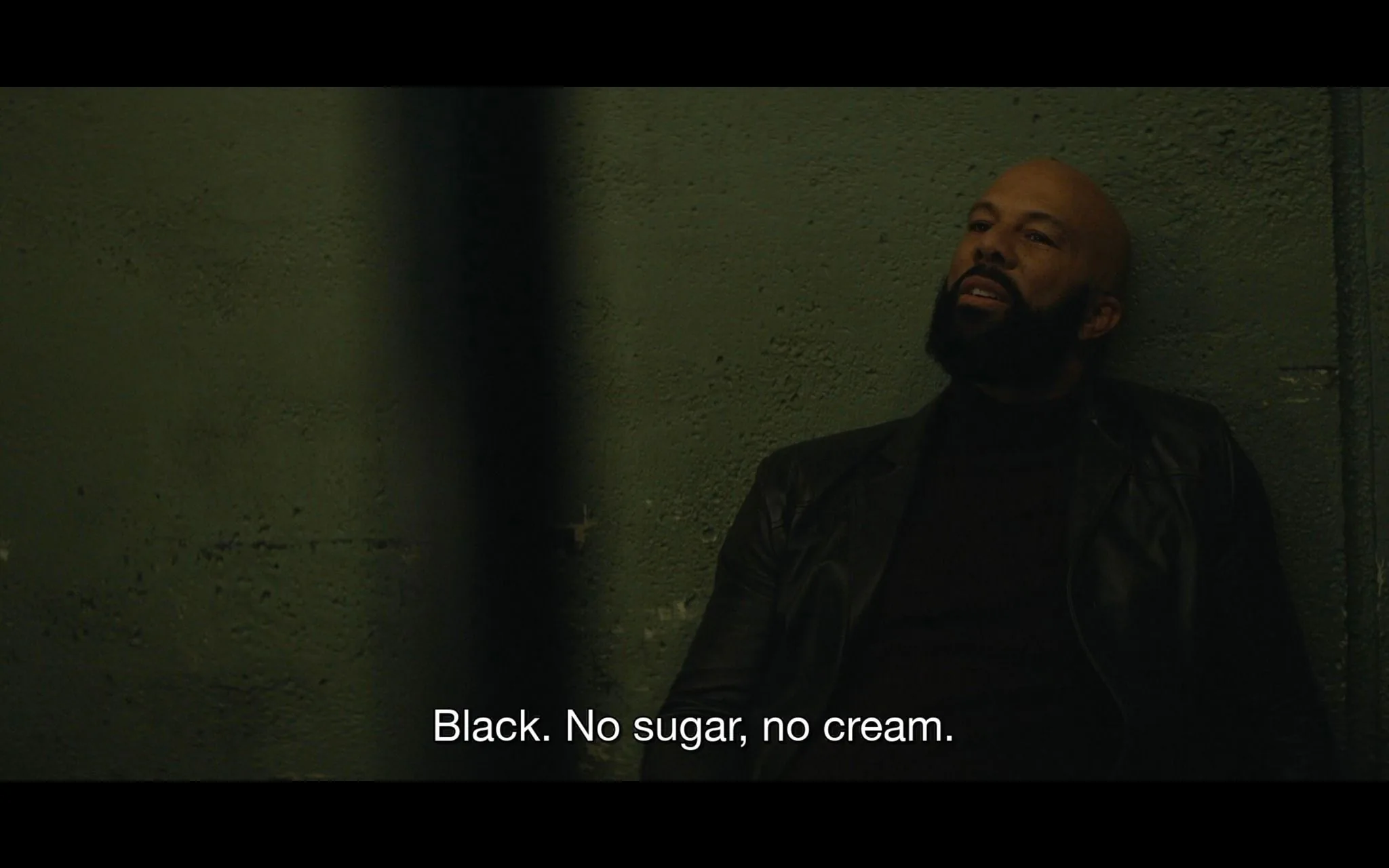 The image captures a dimly lit indoor scene where a man is sitting against a textured wall, suggesting a gritty or tense atmosphere. He has a contemplative expression, wearing a dark turtleneck and a leather jacket, which adds to the seriousness of the mood. The lighting is low, casting shadows and giving the scene a somber, intimate feel. The background is plain with a rough surface, likely indicating a utilitarian or industrial setting.
The composition focuses on the man, who seems to be engaged in a conversation, as indicated by the subtitle at the bottom of the frame: "Black. No sugar, no cream." This suggests a dialogue related to a coffee order, highlighting a preference for simplicity. The framing is tight, mostly centered on the man's upper body, enhancing the focus on his facial expression and attire. This scene appears to be from a film or series, characterized by its moody lighting and minimalistic setting.