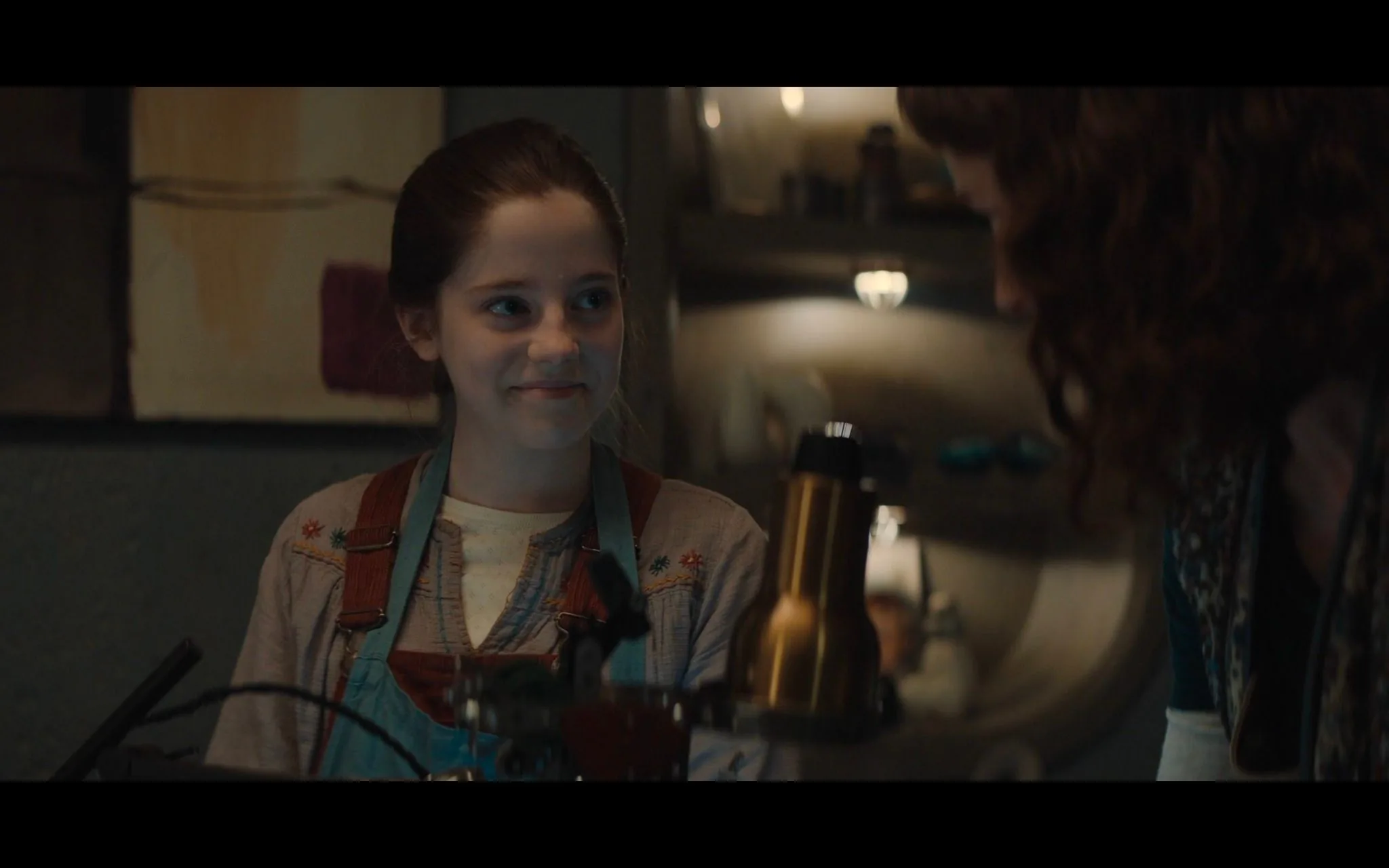 The image is a scene from "Ahsoka," featuring a young girl in a workshop setting. She has her hair pulled back and is wearing overalls with a decorative shirt underneath. Her expression suggests a sense of curiosity or amusement as she interacts with another person in the frame, whose face is not visible. The lighting in the room is warm, casting a soft glow over the scene. In the foreground, there are various tools and equipment, suggesting a focus on mechanical work or tinkering. The background shows shelves with miscellaneous items, adding depth to the workshop environment. The overall atmosphere is one of focus and collaboration, typical of a setting where ideas and inventions come to life.