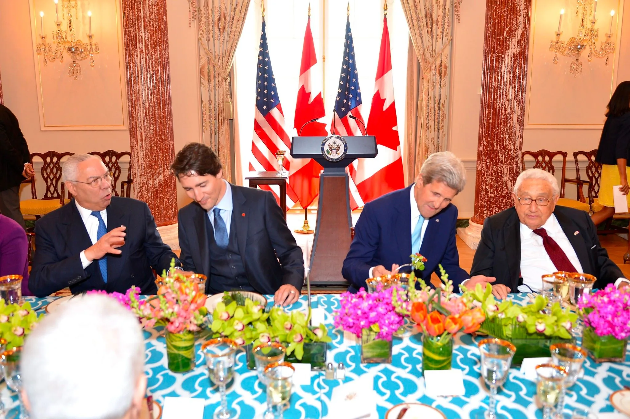```
This image captures a formal meeting setting with notable political figures. Seated at a table adorned with a vibrant blue and white tablecloth and floral centerpieces, the atmosphere is both elegant and diplomatic. From left to right, Colin Powell is engaged in conversation, gesturing expressively. He is dressed in a dark suit with a light blue tie, exuding a sense of authority and engagement.
Next to him, Justin Trudeau is listening attentively, leaning slightly forward. He is dressed in a dark suit with a vest and a patterned tie, displaying a demeanor of interest and respect. John Kerry, beside Trudeau, is focusing on his plate, engaging in the meal. He is wearing a dark navy suit with a blue tie, maintaining a poised and composed appearance.
Finally, Henry Kissinger sits to the right, observing the proceedings. He is in a dark suit with a white shirt and maroon tie, portraying an image of wisdom and experience. In the background, there are U.S. and Canadian flags, and a podium with the seal of the United States, indicating a diplomatic event. The room is richly decorated, with chandeliers and ornate curtains, enhancing the formal and official ambiance.