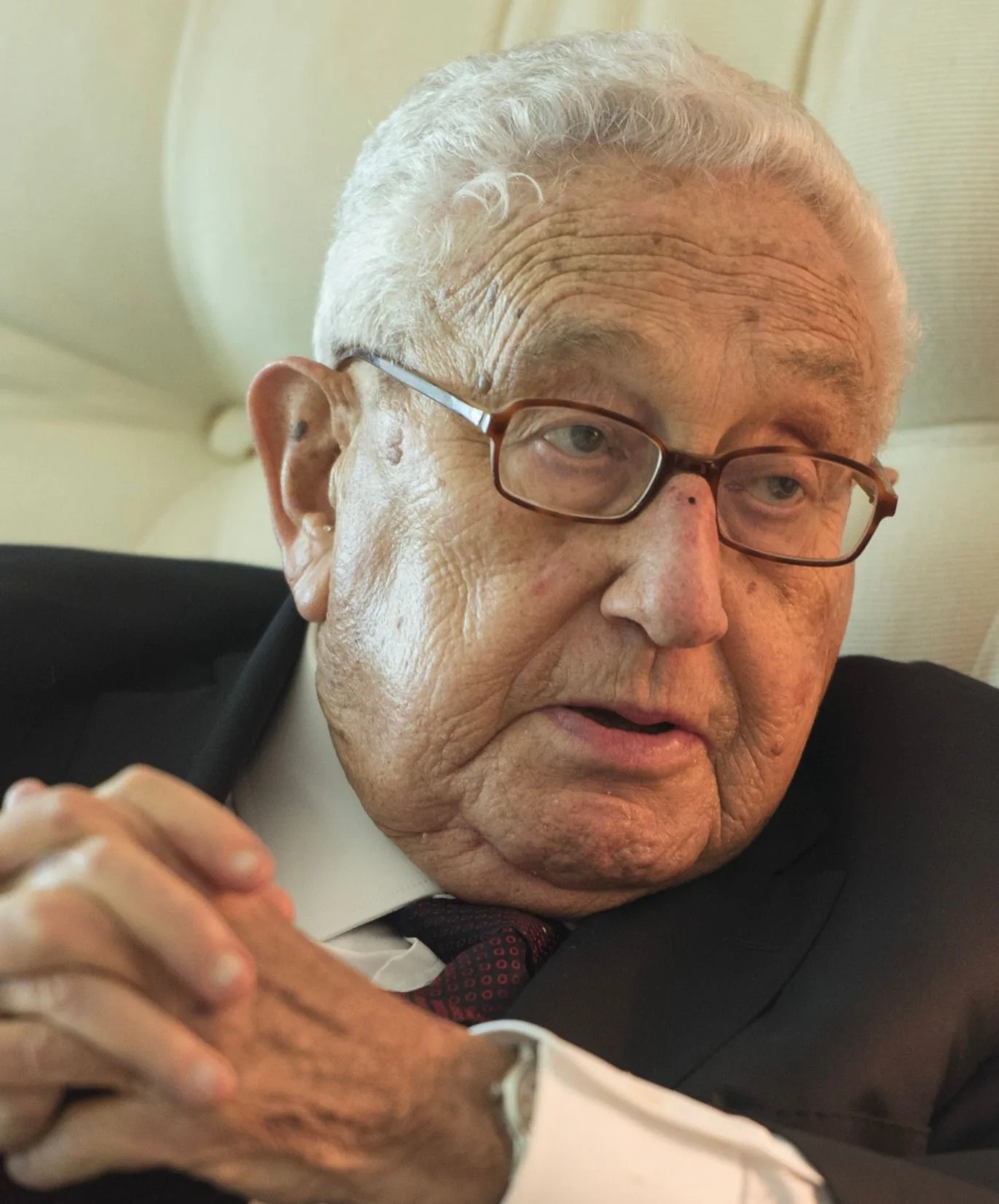 Henry Kissinger is depicted in this image. He is seated indoors, likely in a formal setting, given the upholstered background that suggests a plush chair or sofa. Kissinger is wearing a dark suit, a white dress shirt, and a patterned tie, which denotes a professional and distinguished appearance. His hands are clasped together thoughtfully, suggesting contemplation or engagement in a conversation. Kissinger's expression is attentive and serious, with his eyes directed slightly to the side. His glasses have a light brown frame, complementing his overall attire. The lighting is soft, highlighting the texture of his skin and the details of his face, conveying a sense of warmth and calm. The composition focuses closely on his upper body and face, creating an intimate and personal portrait.