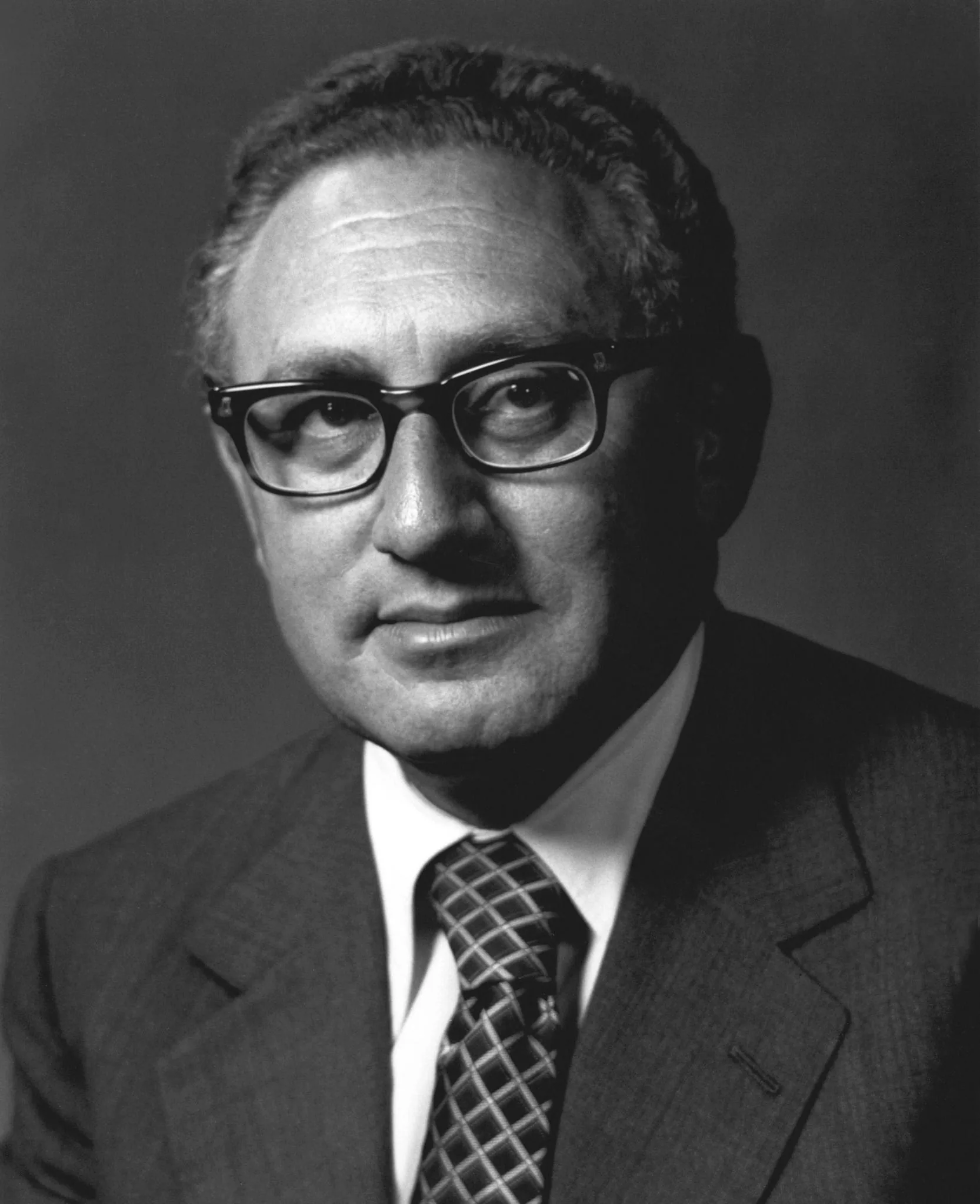 Henry Kissinger is depicted in this black-and-white portrait. The image captures him in a formal setting, where he is wearing a suit and tie, indicative of his professional and diplomatic roles. His suit is dark, complementing the intricately patterned tie that adds a touch of personality to his attire. Kissinger's expression is composed and serious, reflecting the gravitas often associated with his work in international relations. His eyeglasses are prominent, highlighting his intellectual persona. The background is plain and unobtrusive, ensuring that the focus remains solely on him. The lighting is soft, casting gentle shadows that enhance the depth of the image. This portrait conveys a timeless quality, characteristic of official photographs meant to capture influential figures at pivotal moments in their careers.