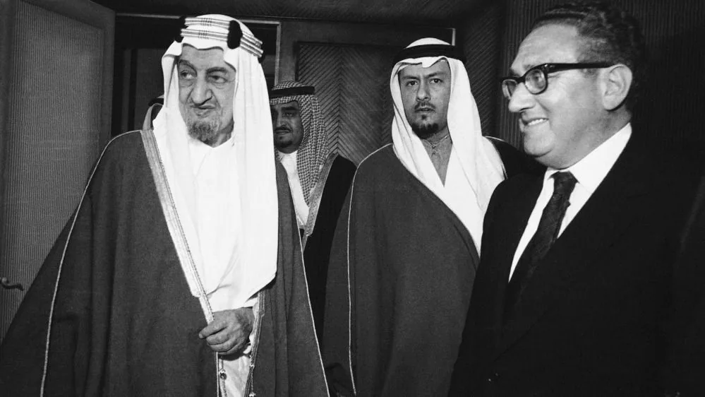 ```
The black-and-white image captures a formal and historically significant meeting. To the right, Henry Kissinger is visible, smiling, wearing a dark suit and tie. His demeanor appears confident and engaged. The individuals with him are dressed in traditional Middle Eastern attire, including white robes and head coverings, indicative of their cultural background. The setting seems to be indoors, likely a conference or meeting room, suggested by the formal attire and serious expressions of the individuals.
The others in the image are dressed in long robes and headscarves, signifying high-ranking officials or royalty from a Middle Eastern country. Their expressions are serious and focused, contrasting with Kissinger's smile. The lighting in the room is even, highlighting the details of their clothing and expressions, and the composition emphasizes the interaction and engagement among the figures. The image suggests a moment of diplomatic importance, potentially discussing international relations or agreements.
No visible text or branding is present in the image.