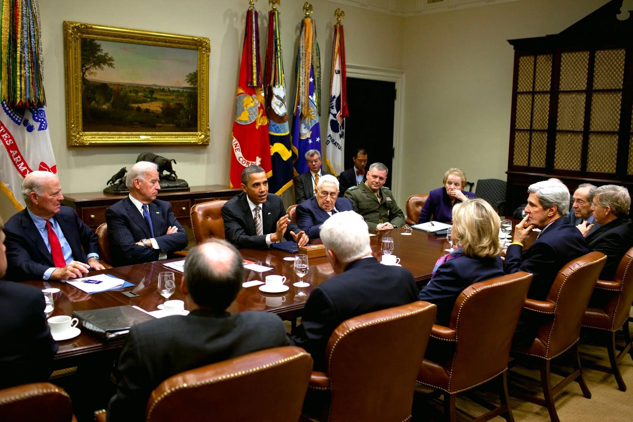 The image depicts a high-level meeting in a conference room, featuring several prominent figures. Barack Obama is visibly speaking, gesturing with his hand to emphasize a point. Seated next to him is Joe Biden, listening intently with his hands clasped. Henry Kissinger is also present, appearing engaged in the discussion.
The room is formal, with a large wooden table surrounded by brown leather chairs. Flags representing various military branches hang in the background, adding to the official atmosphere. The walls are adorned with a large painting and a display of medals or ribbons. John Kerry is seen on the right side, deep in thought, with his hand on his chin. A woman, likely Hillary Clinton, is seated with her back to the camera, identifiable by her blonde hair and attire.
The attendees are dressed in business and military attire, reflecting the serious nature of the meeting. The lighting is bright, illuminating the expressions and gestures of the participants. The mood appears to be focused and collaborative, indicative of a strategic or diplomatic discussion.