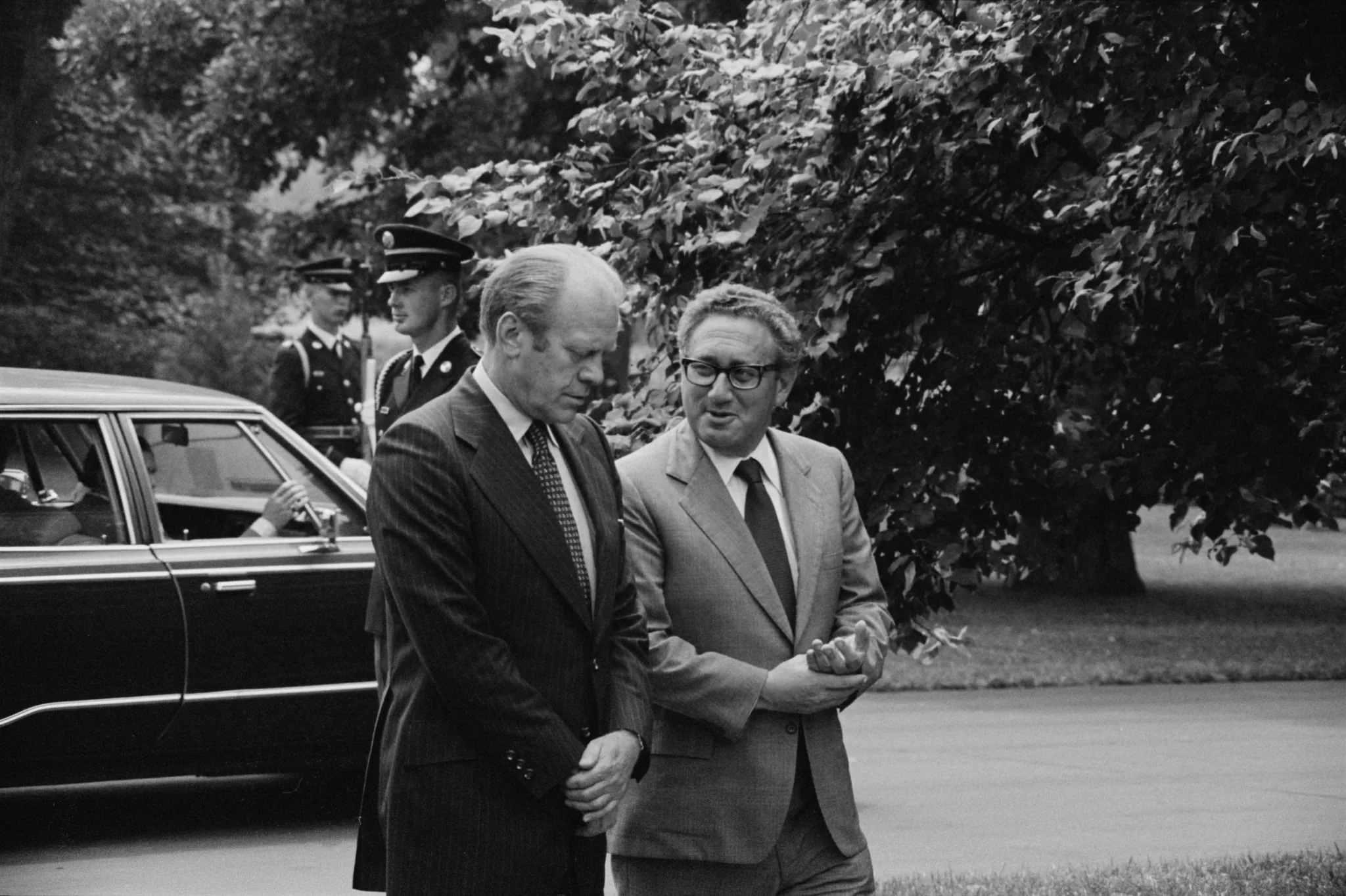 ```
In this black and white photograph, two men, identified as Gerald Ford and Kissinger, are walking together outside. Gerald Ford, wearing a dark pinstripe suit with a tie, appears to be listening intently with a serious expression. Kissinger, dressed in a lighter suit and tie, seems to be speaking, gesturing with his hands, conveying a sense of engagement in their conversation. They are on a paved path, surrounded by lush greenery, indicating a formal outdoor setting, possibly at a government or official residence.
In the background, a car is partially visible with a person inside, and two uniformed guards stand near the vehicle, adding to the formal atmosphere. The scene captures a moment of serious discussion or consultation between the two figures. The photograph has a historical and candid feel, capturing a slice of political life. The lighting is natural, consistent with an outdoor setting, highlighting the figures against the darker backdrop of the foliage.