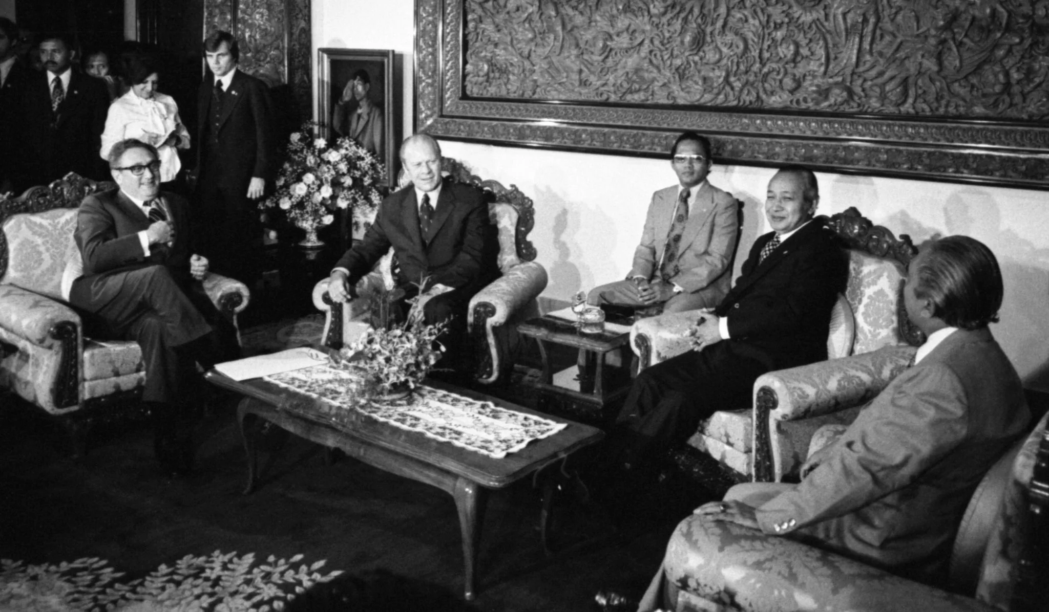 ```
The image captures a formal meeting setting with several individuals seated in an elegantly furnished room. In the center, two individuals are seated on ornate armchairs with a decorative wooden table between them, adorned with a floral arrangement and a lace runner. The person on the left, according to the hint, is Henry Kissinger. He is wearing a dark suit and glasses, and appears to be engaged in conversation with others in the room. Seated beside him is another man in a suit, likely participating in the discussion.
The room is richly decorated, featuring an intricate tapestry hanging on the wall behind the group and elegant floral arrangements. The atmosphere suggests a diplomatic or official setting, with people dressed in formal attire. There are additional individuals in the background, some standing near a doorway, attentively observing the interaction. The lighting is bright, illuminating the faces of those present and highlighting the luxurious details of the room's décor.