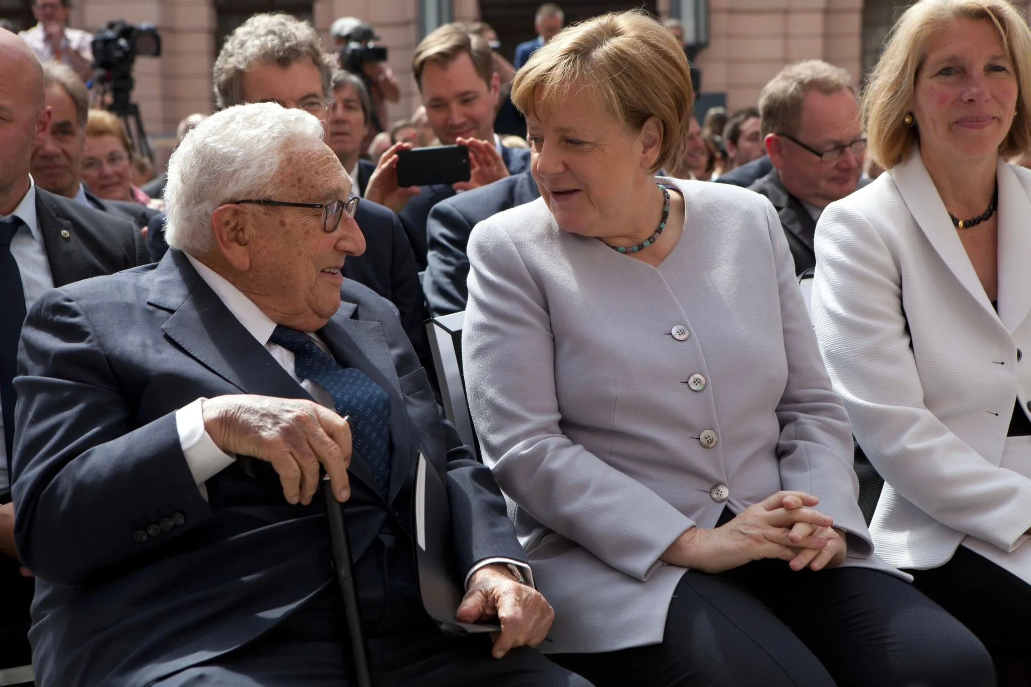 ```
The image captures a moment between Henry Kissinger and Angela Merkel at an outdoor event. They are seated next to each other, engaged in conversation. Henry Kissinger, on the left, is dressed in a dark suit with a white shirt and patterned tie, holding a cane. Angela Merkel, on the right, is wearing a light-colored blazer with matching buttons and a beaded necklace. The mood appears cordial and engaged, with Merkel leaning slightly towards Kissinger as they converse.
The setting is a public gathering, likely formal, suggested by the attire of those present and the presence of photographers and onlookers in the background. The lighting is natural, indicating that the event is taking place during the day. The background shows several people seated in rows, with some using cameras or phones to capture the scene. Overall, the image conveys a moment of interaction between two notable figures in a formal, public setting.