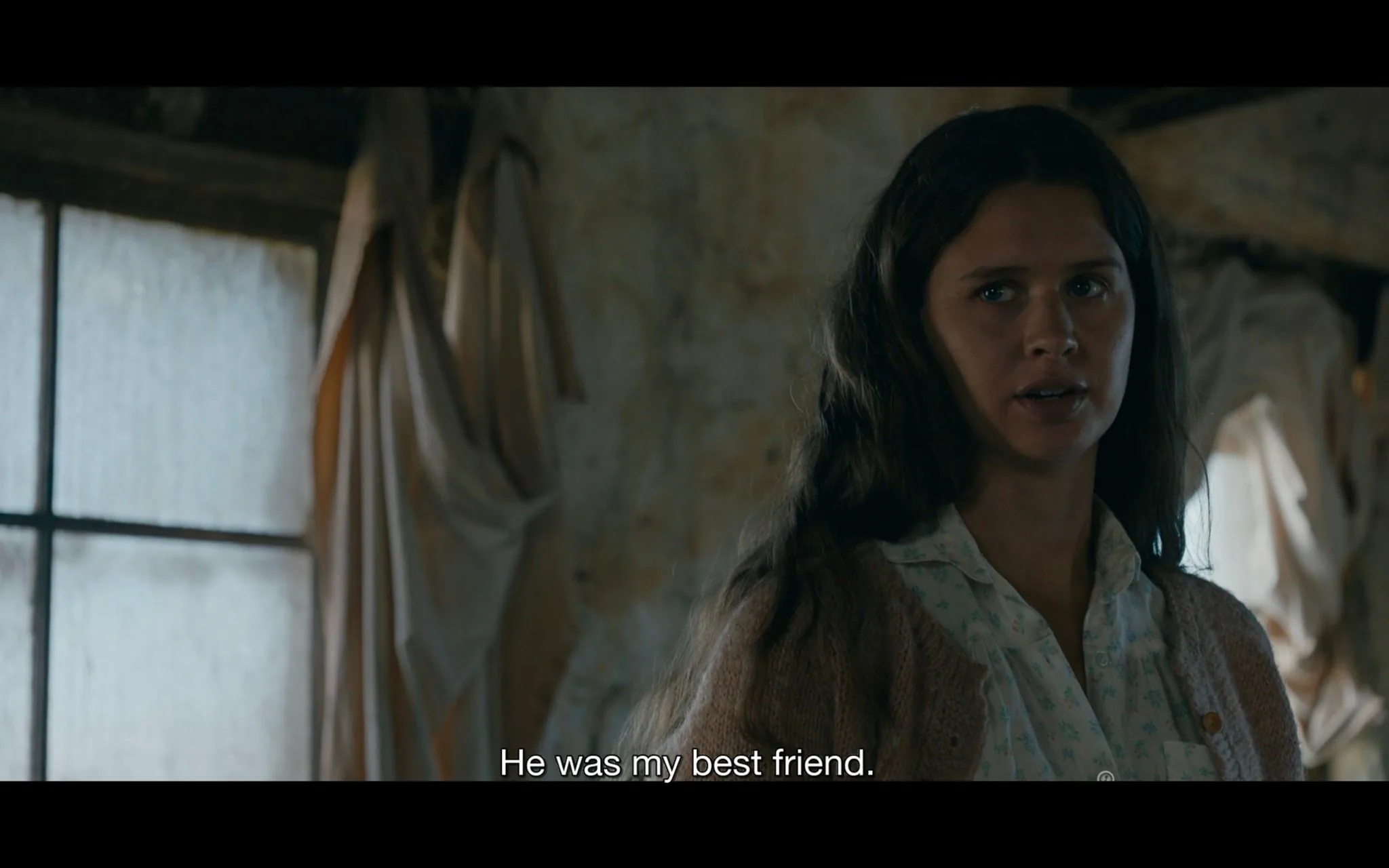 The image is a screenshot from the TV show "from," produced by MGM. It features a young woman indoors, likely part of a dramatic scene. She is positioned slightly off-center, looking towards someone or something outside the frame with a serious expression. Her long, dark hair is naturally styled, and she wears a light-colored blouse with a patterned cardigan. The background shows a rustic, somewhat worn interior with a window partially covered by draped fabric. The lighting is soft but dim, creating an intimate and somber atmosphere. The subtitle at the bottom of the image reads, "He was my best friend," indicating an emotional context. The scene suggests a moment of reflection or revelation, enhanced by the muted colors and the character's earnest demeanor.