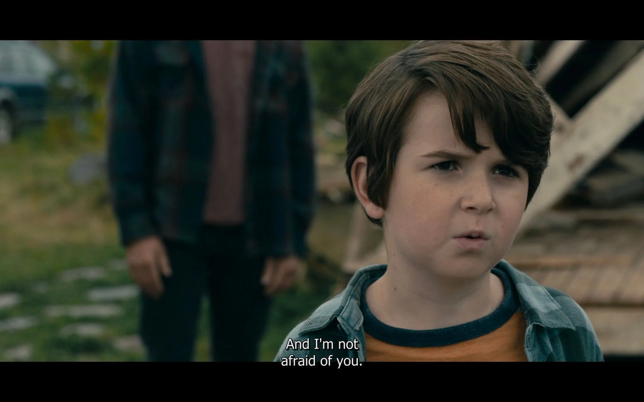 The screenshot is from the TV show "From" by MGM. It depicts a tense outdoor scene during the daytime. In the foreground, a young boy stands facing forward, displaying a determined expression. He wears a plaid shirt over a mustard-colored t-shirt, and his hair is neatly styled. The background is slightly blurred, showing someone standing behind him, out of focus, possibly an adult, suggesting a confrontation or a moment of defiance.
The setting appears rustic or rural, with some wooden structures and grassy areas visible. The lighting is natural, indicating it is daytime. The composition centers on the boy, emphasizing his bold stance and the seriousness of the moment. The subtitle at the bottom of the frame reads, "And I'm not afraid of you," highlighting the boy's courage or defiance in the scene. The mood is tense, and the visual focus remains primarily on the boy's expression and stance.