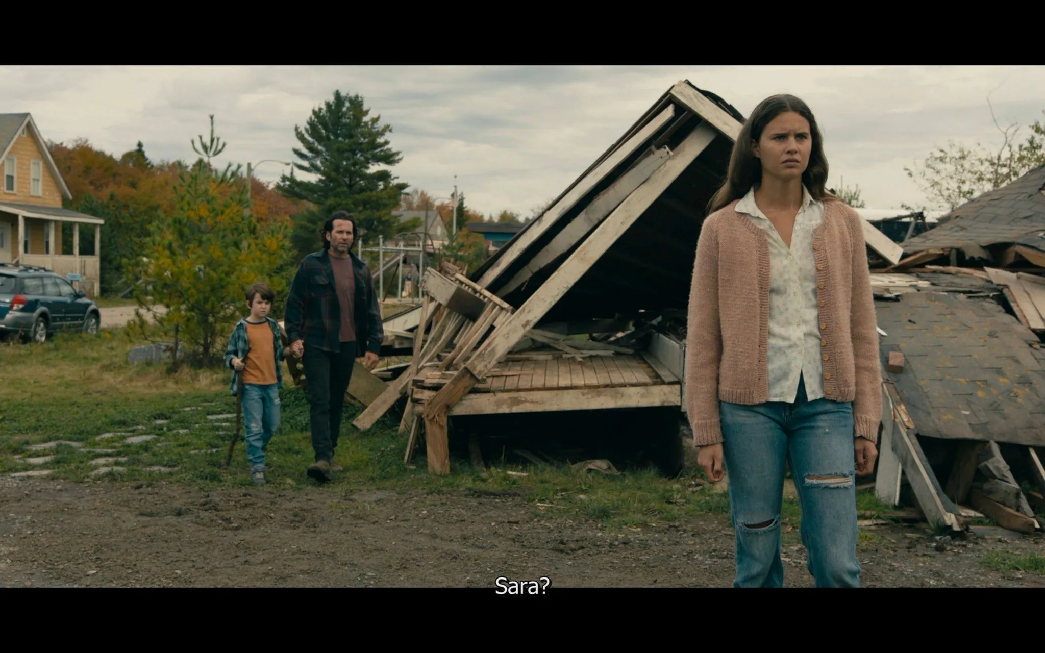 The image is a screenshot from the TV show "From." In the foreground, a young woman stands with a concerned expression. She is wearing a light-colored blouse and a pink cardigan, paired with ripped jeans. Her posture suggests she might be confronting or processing something significant. Behind her, there is a collapsed wooden structure, possibly a shed or house, indicating some form of destruction or decay.
In the background, a man and a child walk towards her. The man wears a dark plaid jacket and the child is dressed casually, holding his hand. The setting is outdoors with a rural or suburban atmosphere, featuring a grassy area and a parked vehicle in front of a house. The weather appears overcast, casting a muted light over the scene, enhancing the somber mood. The subtitle reads "Sara?", suggesting a moment of recognition or inquiry about the woman in the foreground.
