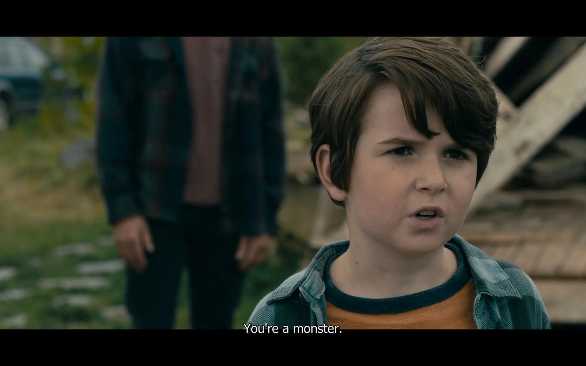 The image is a frame from the TV show "From" by MGM. It depicts a young boy in an outdoor setting, likely in a dramatic or tense moment, as his expression is serious and focused. He is wearing a blue plaid shirt over an orange T-shirt, standing in front of a blurred background that includes some wooden structures and greenery. The scene captures a confrontation, emphasized by the subtitle "You're a monster," suggesting a moment of emotional intensity. The lighting is natural, indicating it is daytime, and the composition centers on the boy, with an adult figure slightly out of focus in the background. The mood appears tense and confrontational, contributing to the dramatic tone of the scene.