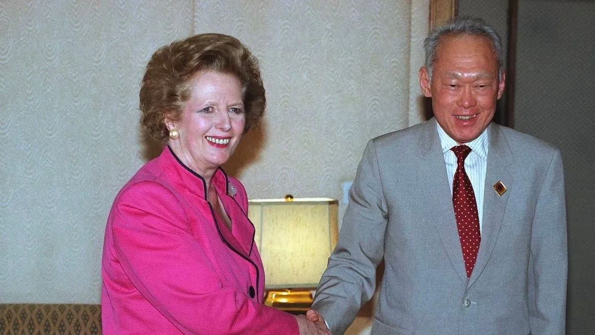 The image captures a diplomatic meeting between Maggie Thatcher and Lee Kuan Yew. They are indoors, possibly in a conference or office setting, characterized by a neutral-toned wallpaper and a table lamp in the background that provides soft lighting. Maggie Thatcher is wearing a bright pink blazer with black piping, which contrasts with the more subdued background. Her hair is styled in a typical 1980s fashion, and she is smiling warmly, indicating a positive and cordial interaction. Lee Kuan Yew is dressed in a light gray suit paired with a white shirt and a red polka-dot tie. He appears to be in good spirits, also smiling as he shakes hands with Maggie Thatcher. Both individuals exude a sense of professionalism and mutual respect, which is highlighted by their formal attire and the setting of their meeting. The overall mood is one of amiability and diplomatic engagement.