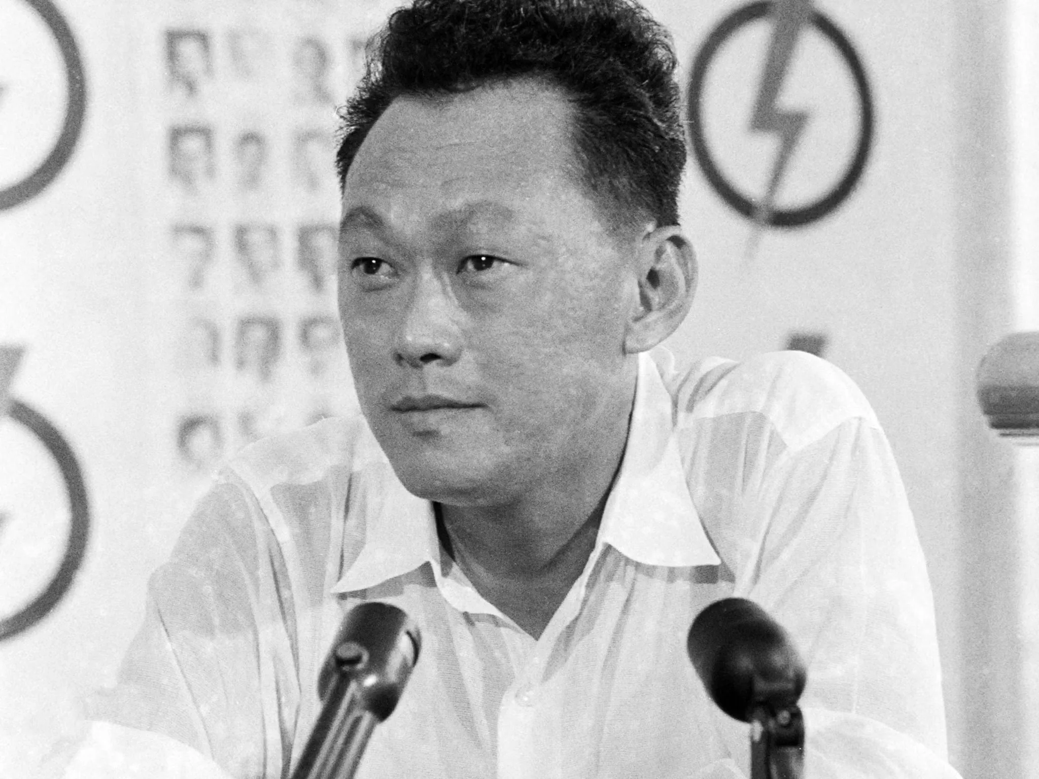Lee Kuan Yew is shown in this black and white image, which appears to be from a press conference or public speaking event. He is positioned centrally, sitting at a table with a microphone in front of him. His attire is a simple, light-colored shirt, which contrasts sharply with the darker microphone and the subtle background elements. The backdrop features some blurred symbols and what appears to be a logo with a lightning bolt, possibly indicating a political or organizational setting. Lee Kuan Yew's expression is focused and contemplative, suggesting he is engaged in a serious discussion or announcement. The lighting is even, typical of indoor environments, without any dramatic shadows, emphasizing the straightforward, documentary style of the photograph. The setting conveys a sense of historical gravitas and political significance.