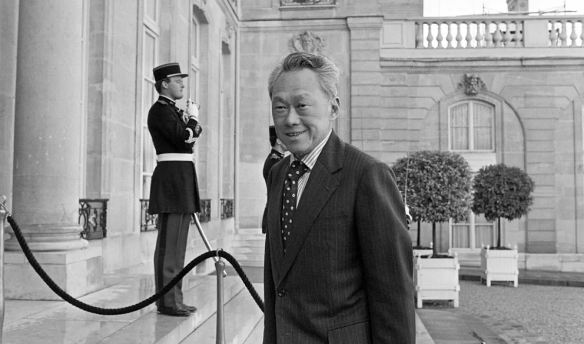 Lee Kuan Yew is captured in this historical black and white photograph. He stands in front of an elegant building, possibly a significant government or diplomatic location, as indicated by the architecture and presence of a uniformed guard. The guard, positioned to the left, wears a formal uniform with a hat and appears to be standing at attention. Lee Kuan Yew is dressed in a well-tailored, striped suit with a polka dot tie, exuding an air of formality and authority. His expression is calm and confident, reflecting the demeanor of a prominent statesman. The setting is outdoor, with neatly trimmed potted trees lining the entrance, emphasizing the grandeur of the location. The photograph, composed with Lee Kuan Yew prominently in the foreground, captures a moment of significance, possibly during a diplomatic visit or meeting. The black and white medium adds a timeless quality to the image, highlighting its historical importance.