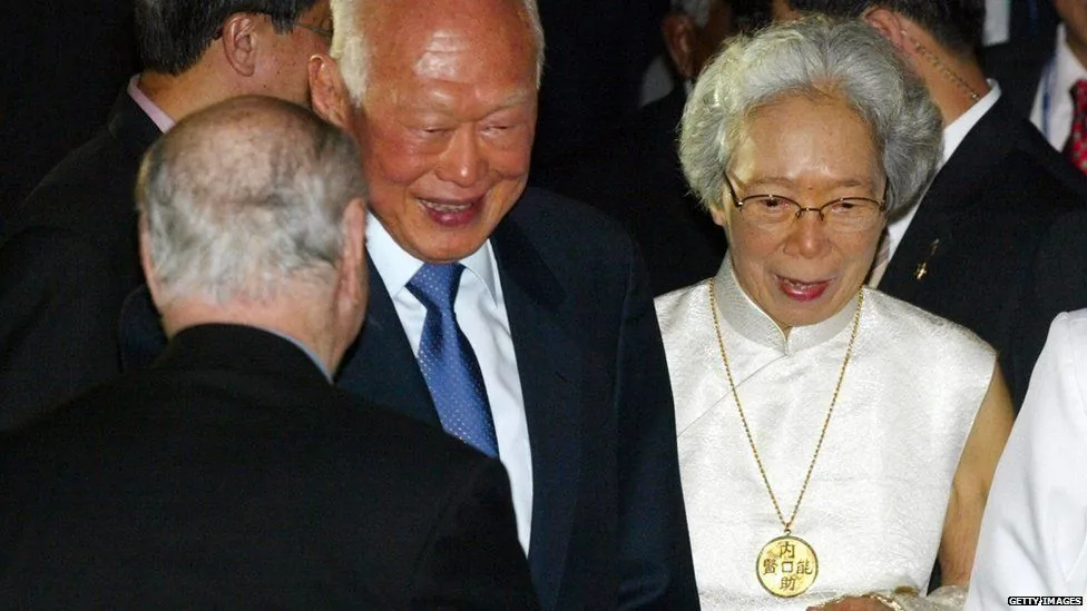 Lee Kuan Yew is prominently featured in this image. He is wearing a dark suit with a white shirt and a blue tie, appearing in a formal setting, likely at a diplomatic or political event. He is smiling, suggesting a cordial atmosphere. Beside him is a woman dressed in a white traditional outfit with a large circular medallion around her neck, indicating a significant ceremony or gathering. The background includes several other individuals, dressed formally, adding to the sense of an important event. The lighting is subdued, emphasizing the formal nature of the occasion. The overall mood of the image is respectful and celebratory, with interactions between the attendees marked by smiles and formal attire.