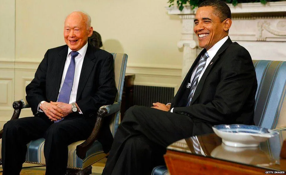 Lee Kuan Yew and Obama are seated in a formal setting, likely a meeting or diplomatic event. They are both smiling, creating a warm and cordial atmosphere. Lee Kuan Yew is wearing a dark suit with a light blue patterned tie, and his hands are resting on his lap. Obama is also dressed in a dark suit, complemented by a light shirt and a striped tie. The room has an elegant decor, featuring a fireplace in the background and a blue-striped armchair. A bowl is placed on the table in front of them, adding to the formal setting. The lighting is bright and evenly distributed, highlighting the expressions and attire of both individuals, contributing to the overall professionalism and warmth of the scene.