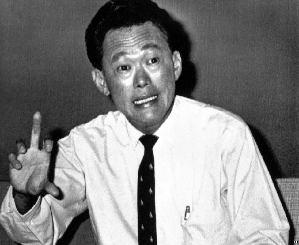Lee Kuan Yew is captured in a black and white photograph, conveying a moment filled with intensity and expression. He is dressed formally in a white shirt and a dark tie with a subtle pattern, suggesting a professional or governmental setting. His facial expression is animated, with wide eyes and an open mouth, as if emphasizing a point or passionately explaining something. His right hand is raised, fingers splayed, adding emphasis to his gesture. The background is plain and devoid of any distracting elements, directing full attention to his expressive demeanor. The high contrast of the black and white image highlights the details of his attire and the emotions conveyed in his face. The overall composition suggests a scene of significant dialogue or debate, characteristic of Lee Kuan Yew’s influential role in political discussions.
