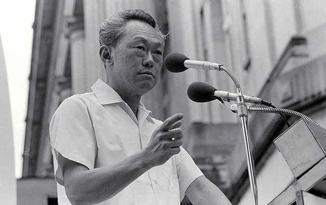 The image is a black-and-white photograph depicting Lee Kuan Yew, known for his significant role in Singapore's history. He stands behind a podium, addressing an audience, with two microphones prominently positioned in front of him. His attire is a simple, short-sleeved white shirt, indicative of a formal yet approachable style. His posture is assertive, with one hand slightly raised, suggesting emphasis is being placed on a point he is making during his speech. The background features a large building with classical architecture, implying the setting might be an important civic location. The lighting is bright, typical of an outdoor event held during the day. His expression is focused and serious, characteristic of someone deeply engaged in delivering an impactful message. Overall, the photograph captures a moment of historical significance, with Lee Kuan Yew at the forefront of leadership and public speaking.