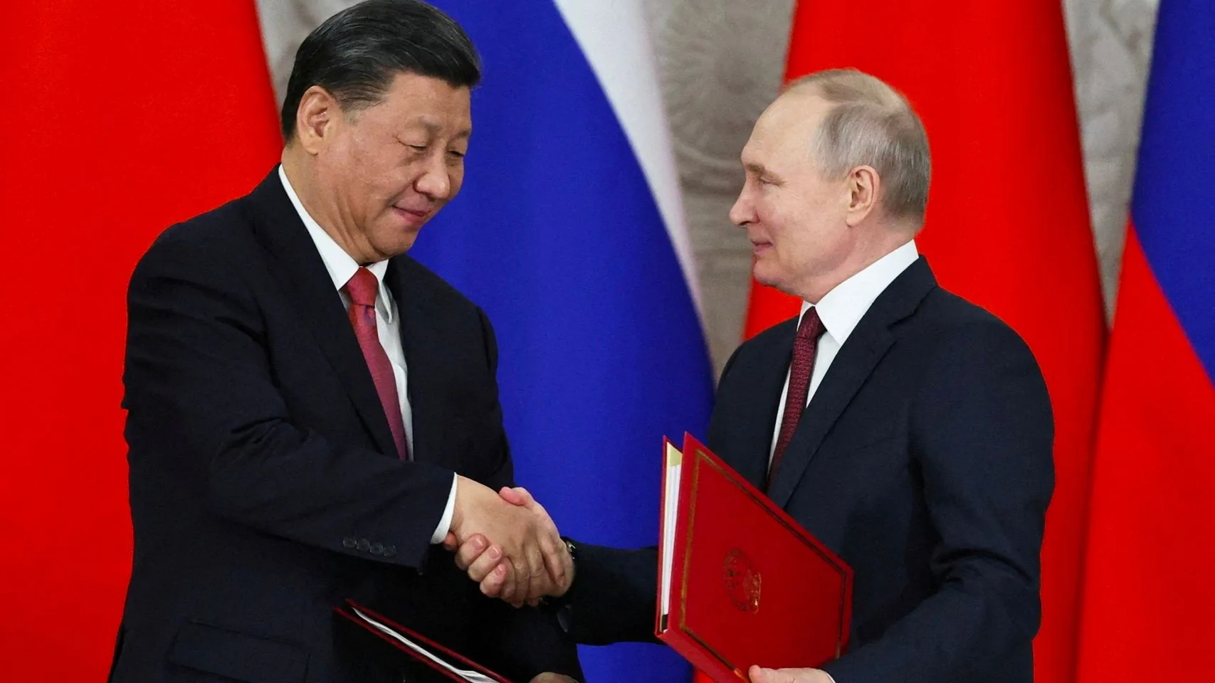 ```
In this image, Xi and Putin are engaging in a formal handshake during what appears to be an official diplomatic ceremony. Both are dressed in dark suits with white shirts and ties, reflecting a formal occasion. Xi is holding a red folder with a subtle pattern on it, while Putin also holds a similar red folder, suggesting the exchange or signing of important documents. Behind them, the background is dominated by large flags, primarily in red and blue, indicating a setting involving their respective countries. The expressions on their faces show a sense of formality and cordiality, underscoring the diplomatic nature of the event. The lighting is bright, highlighting the significance of the moment and ensuring both figures are clearly visible.