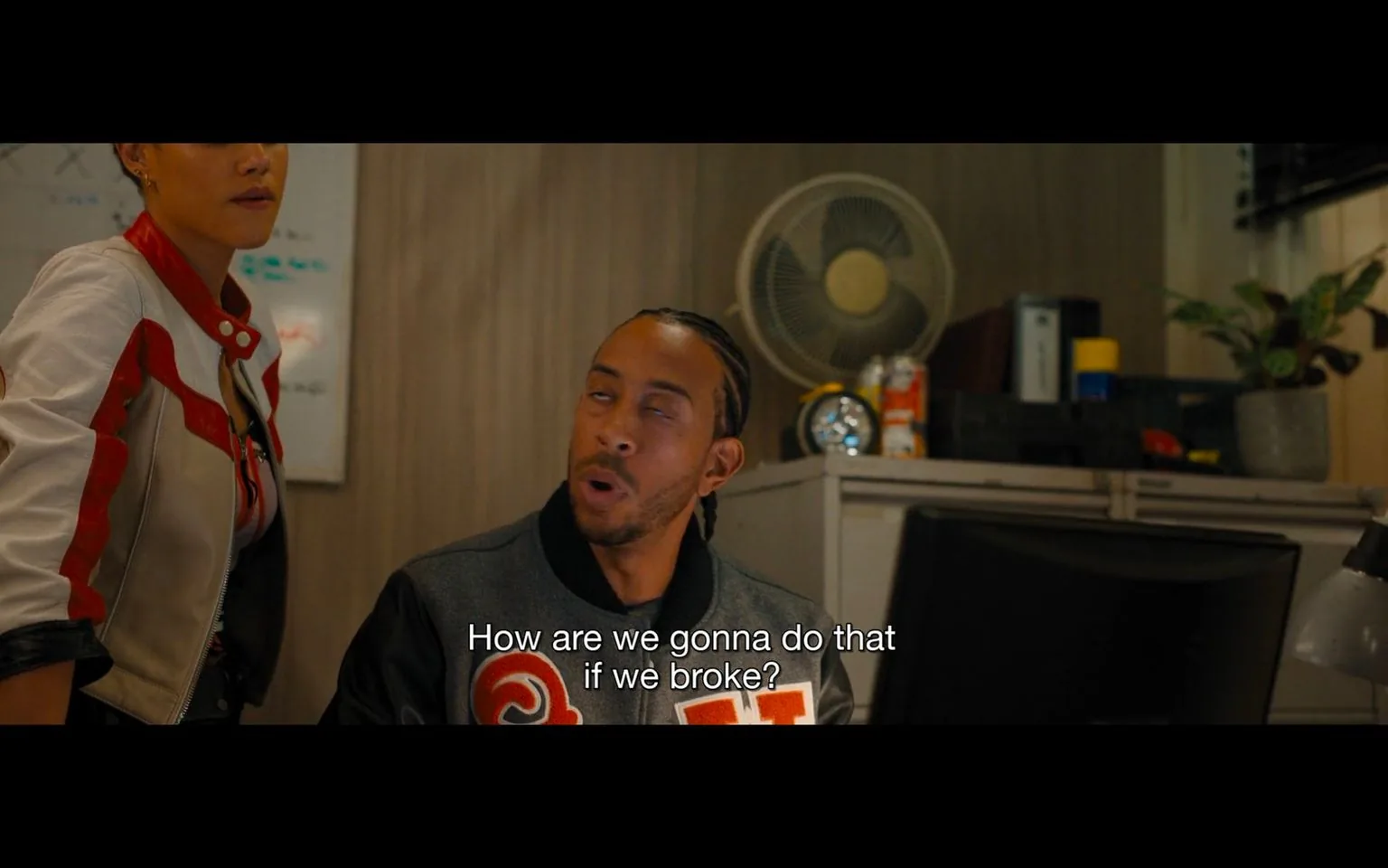 In this scene, the focus is on Ludacris, who appears to be in a conversation, expressing concern with the words, "How are we gonna do that if we broke?" This is set in an indoor office environment, characterized by a desk, a computer monitor, and office supplies scattered around. To the left, a woman is partially visible, suggesting that this is a group discussion. The overall lighting is warm, contributing to a casual and somewhat tense atmosphere. Ludacris is wearing a jacket with a distinctive design, and his expression indicates concern or disbelief. The presence of a fan and some canned drinks in the background adds to the everyday office setting. The composition suggests a planning or problem-solving scene.