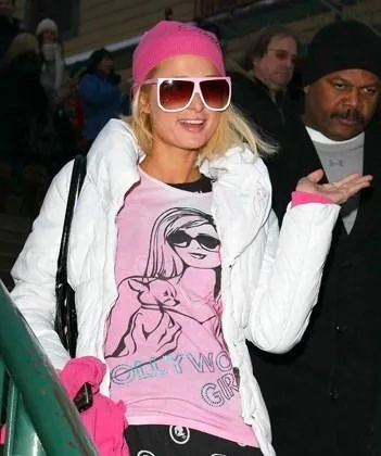 Paris Hilton is pictured outdoors in a casual and fashionable ensemble. She is wearing a bright pink beanie and large white-framed sunglasses that cover much of her face, adding a flashy touch to her look. Her outfit includes a pink graphic t-shirt featuring an illustration of a woman and a small dog with the text "HOLLYWOOD GIRL" visible underneath. Over the t-shirt, she has on a white quilted jacket, suitable for the cold weather suggested by her pink gloves. Hilton's expression appears cheerful and carefree, with her hand raised in a gesturing posture. The background shows other people dressed warmly, indicating a cold day, and the lighting appears to be natural, suggesting it's daytime. The overall vibe of the image is lively and energetic, with Hilton standing out prominently against the more muted background.