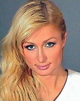 ```
The image features a headshot of Paris Hilton, as identified by the user hint. Paris Hilton is positioned facing the camera with her head slightly tilted, exuding a confident and composed expression. Her long blonde hair cascades over one shoulder, enhancing the focus on her striking blue eyes and subtle makeup that highlights her features. The lighting is even, emphasizing her facial contours against a neutral background typical of a mugshot setting. The image captures a formal portrait style, with Paris wearing a light-colored top that contrasts with the darker colors of the backdrop. The composition is straightforward, focusing solely on her face and upper shoulders, characteristic of a mugshot photograph. The context suggests an official or legal setting, contributing to the serious yet poised demeanor captured in this portrait.