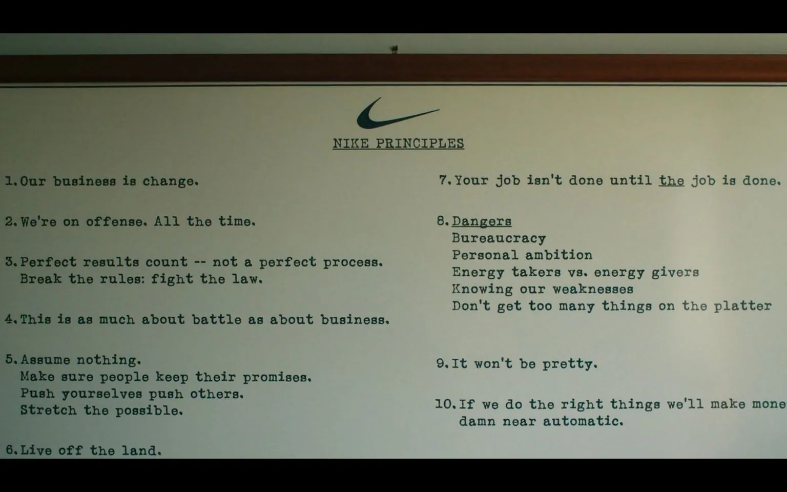 The image shows a framed poster featuring Nike's motivational "Principles." Positioned centrally, the Nike logo is prominently displayed at the top, followed by the title "NIKE PRINCIPLES" in bold, capital letters. The text is organized into a list format, consisting of ten key principles that emphasize themes of change, offense, achieving perfect results, and maintaining a battle-ready mindset in business. These points illustrate Nike’s corporate philosophy, focusing on innovation, perseverance, and strategic thinking. Each principle is succinctly worded, fostering a direct and assertive tone consistent with Nike’s brand identity. The background is a plain off-white, ensuring the text remains the primary focus, with the bold typeface enhancing its readability. The setting appears to be an indoor office space, suggesting a professional environment where such motivational materials would be prominently displayed to inspire employees or visitors. The overall mood conveyed is one of dynamism and determination, reflecting Nike's commitment to excellence and leadership in their field.